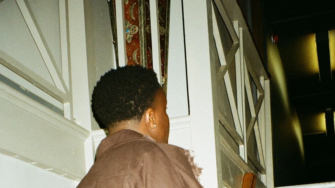 A person wearing a brown jacket stands near a staircase with ornate railing and decorative patterns on a panel. The background is dimly lit.
