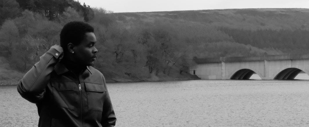 Black and white image of a person in a jacket standing by a body of water with a bridge and forest in the background.