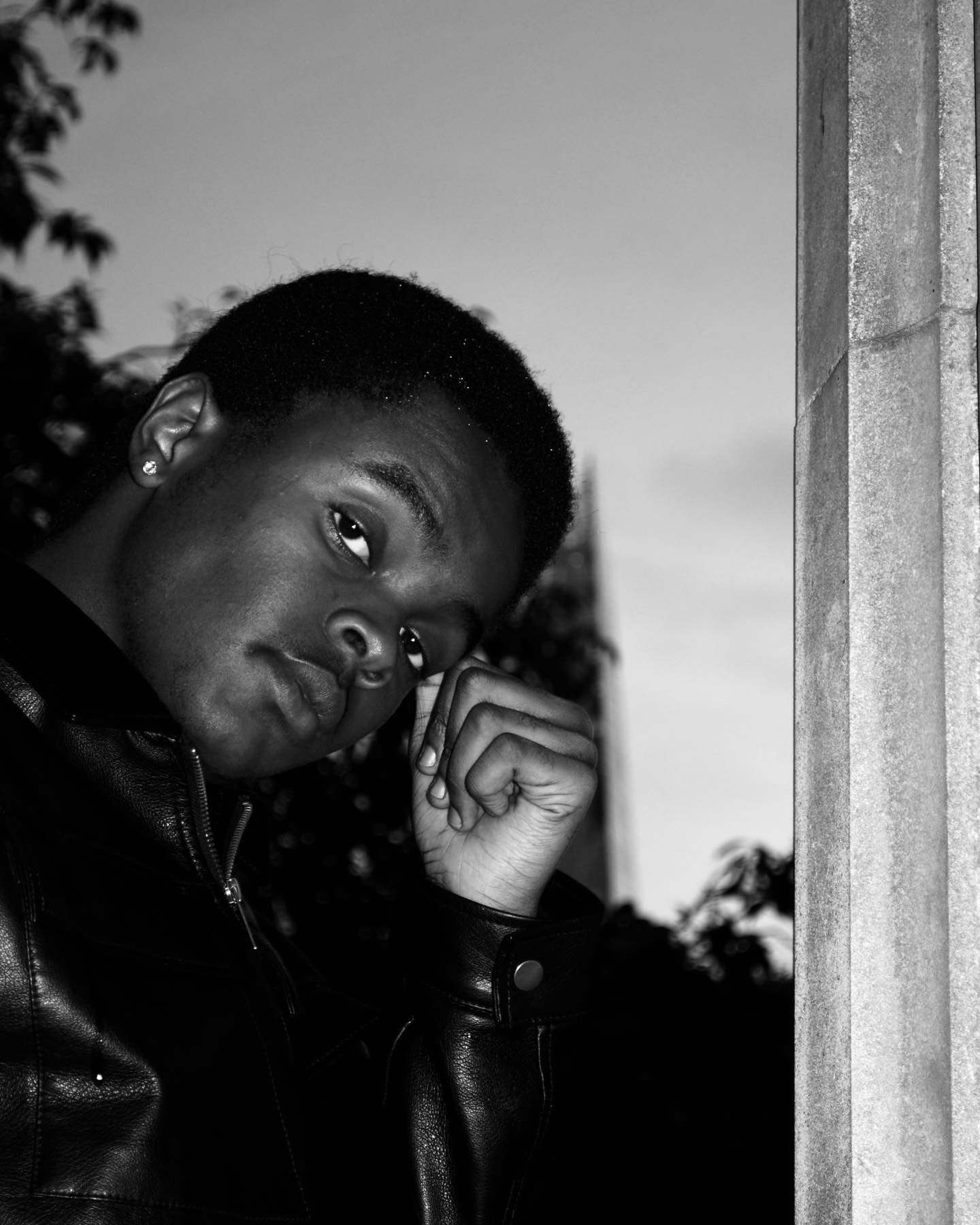 Black and white portrait of a person in a leather jacket leaning against a stone column.