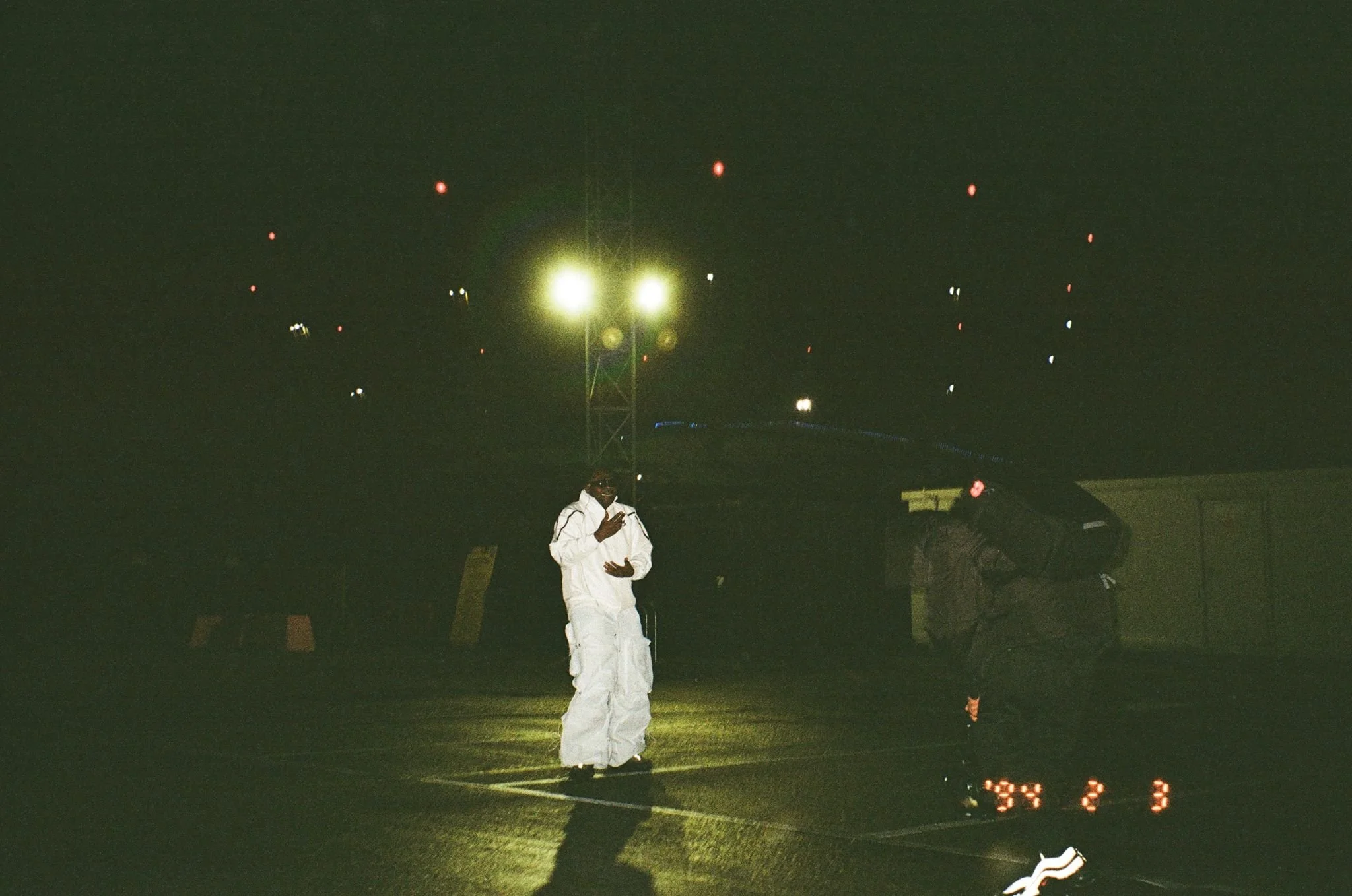 Person in white outfit standing in dimly lit outdoor area with streetlights and distant red lights.
