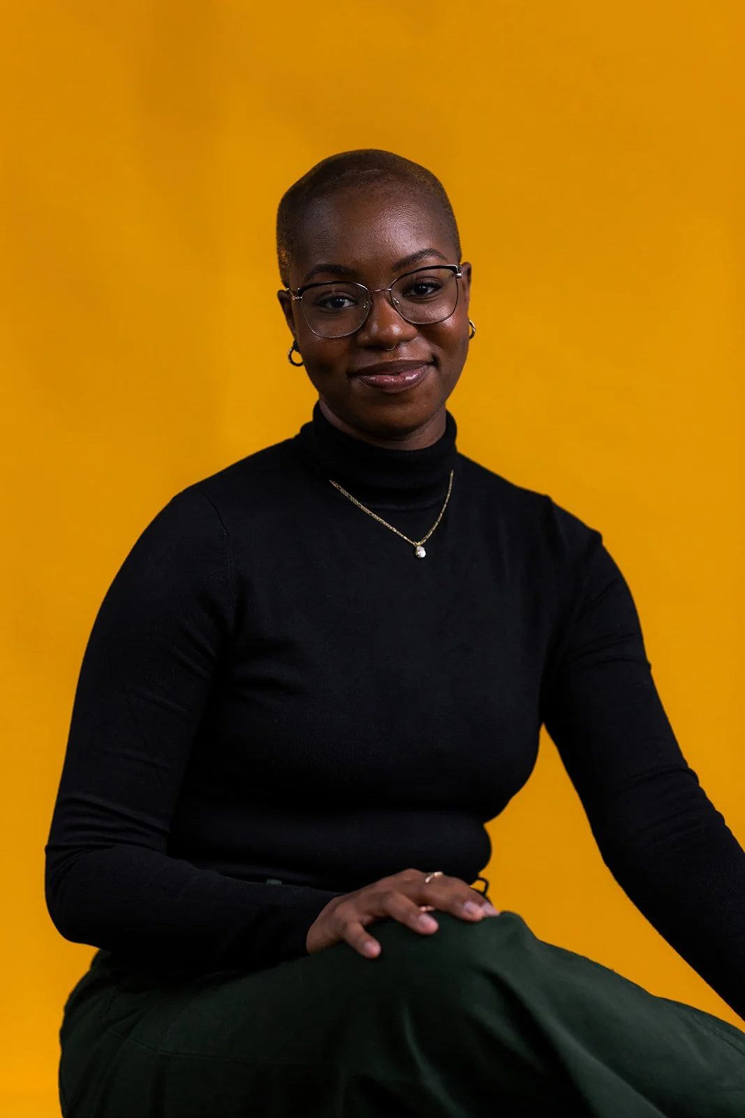 A non-binary person with glasses and a shaved head wearing a black turtleneck and a gold necklace, sitting against a yellow background.