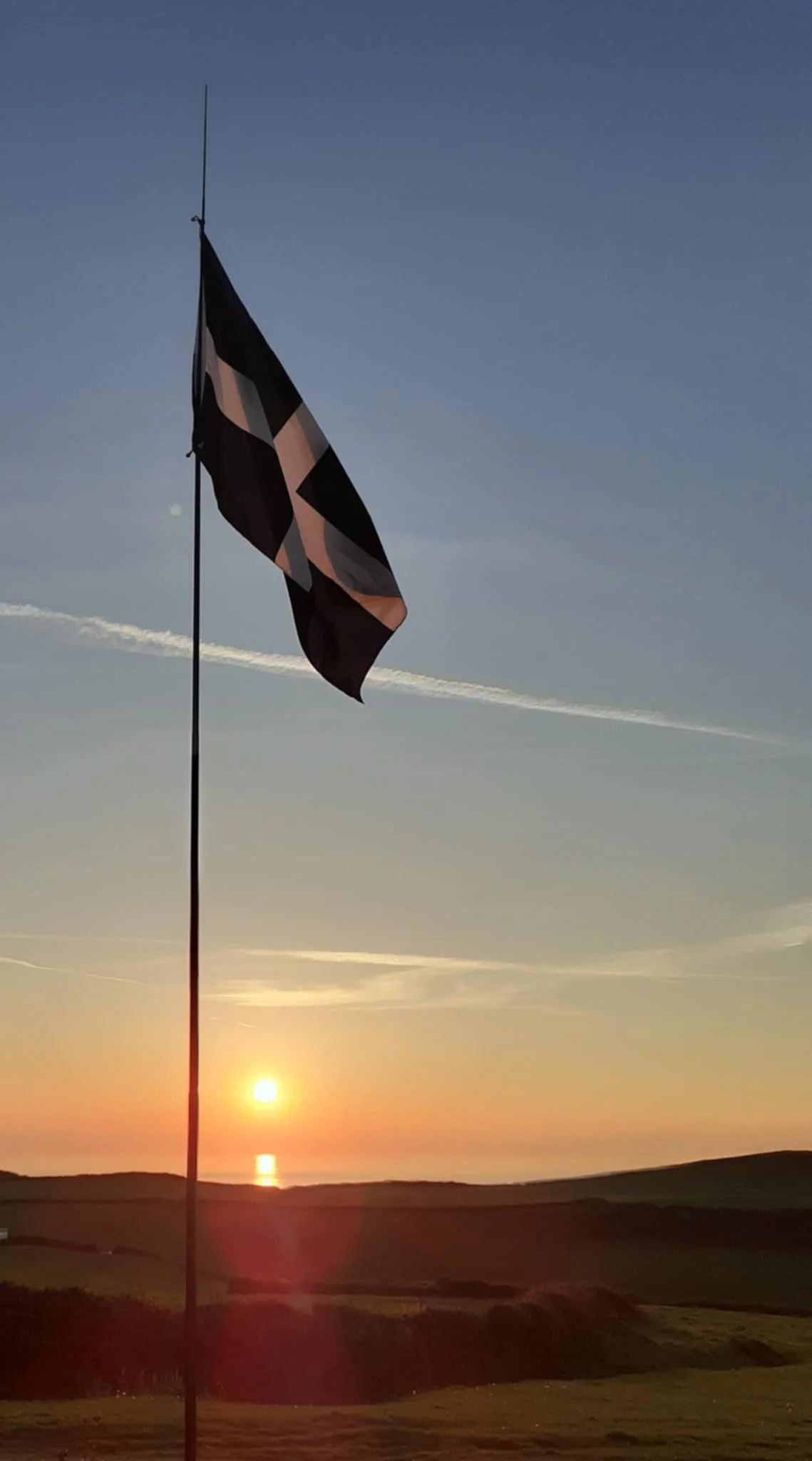 A Cornish flag waving in the wind against a sunset sky with clouds and a distant horizon.