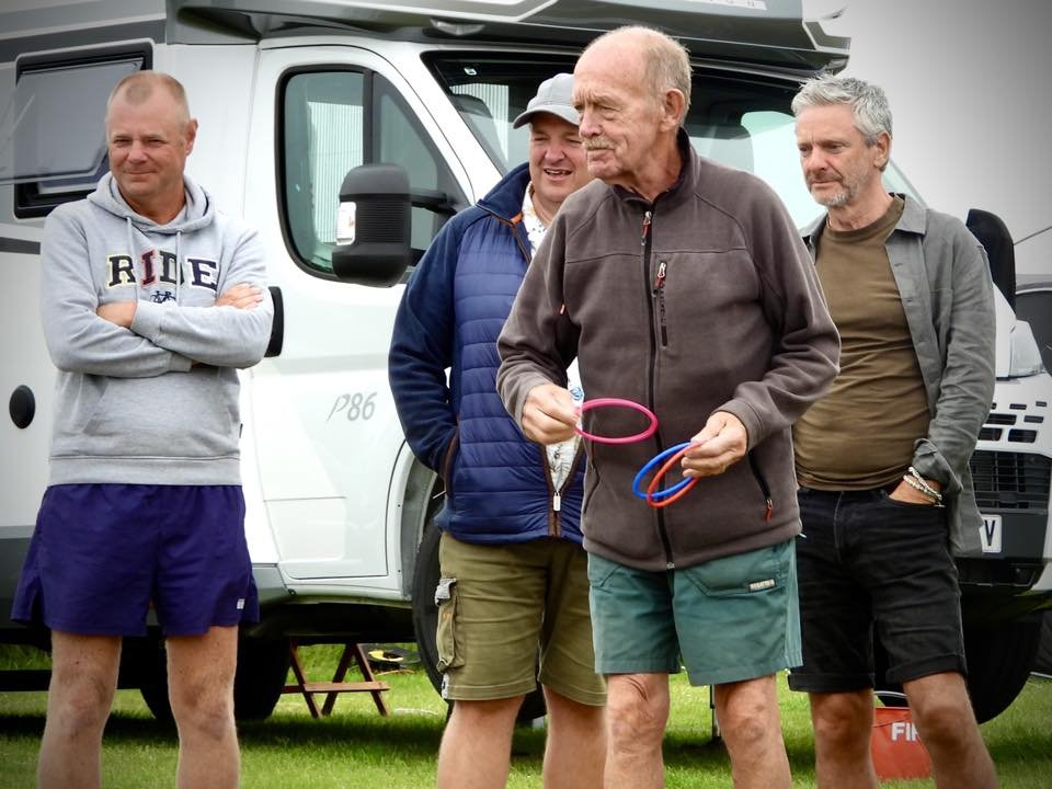 Four men standing outdoors in front of a white camper van. One man in the center is holding rubber rings, possibly for a game or activity. The men are casually dressed, some in shorts and sweatshirts, and are engaged in conversation or observing.