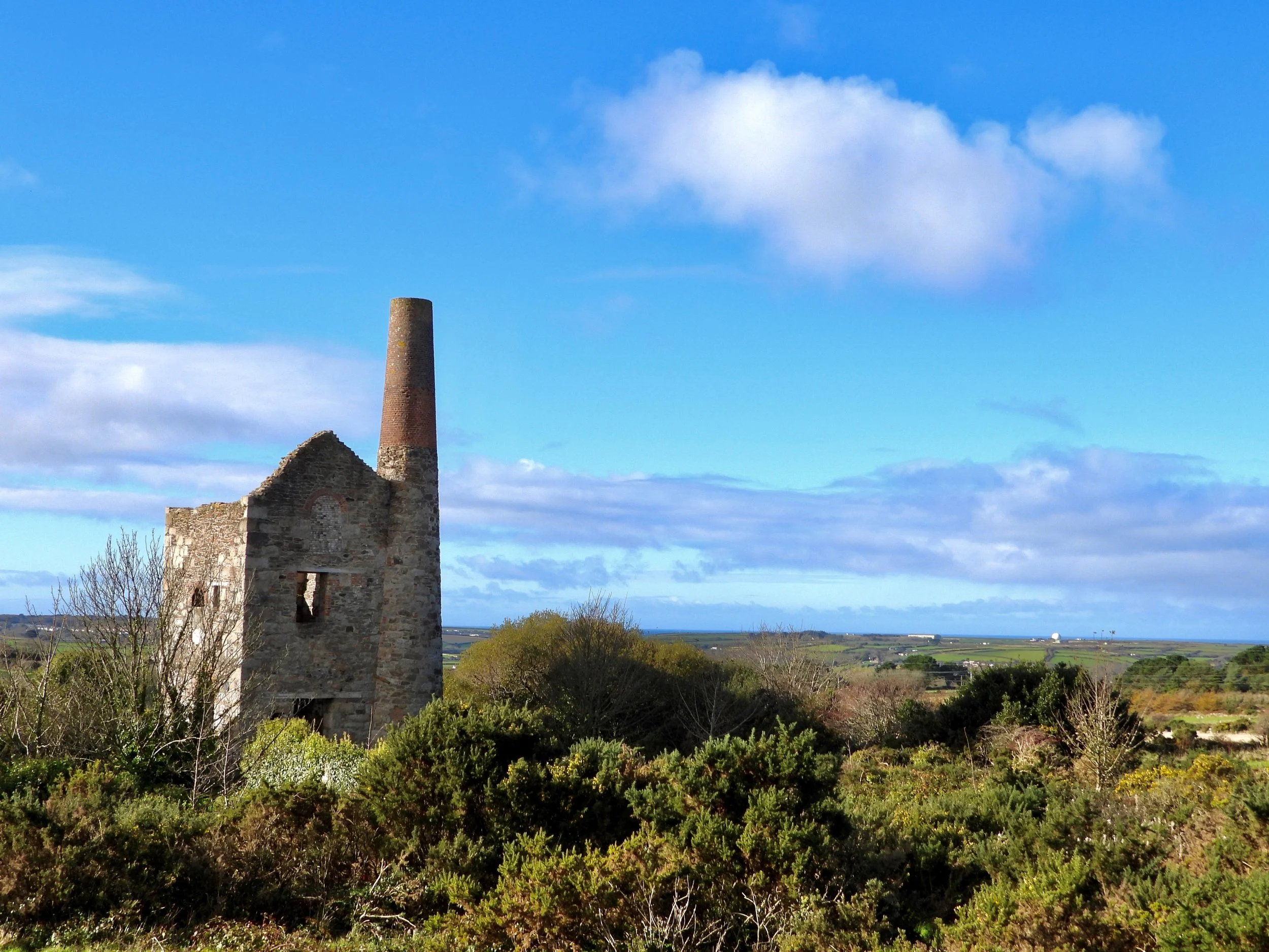 Ruined stone building with a tall chimney surrounded by green bushes under a blue sky with clouds.