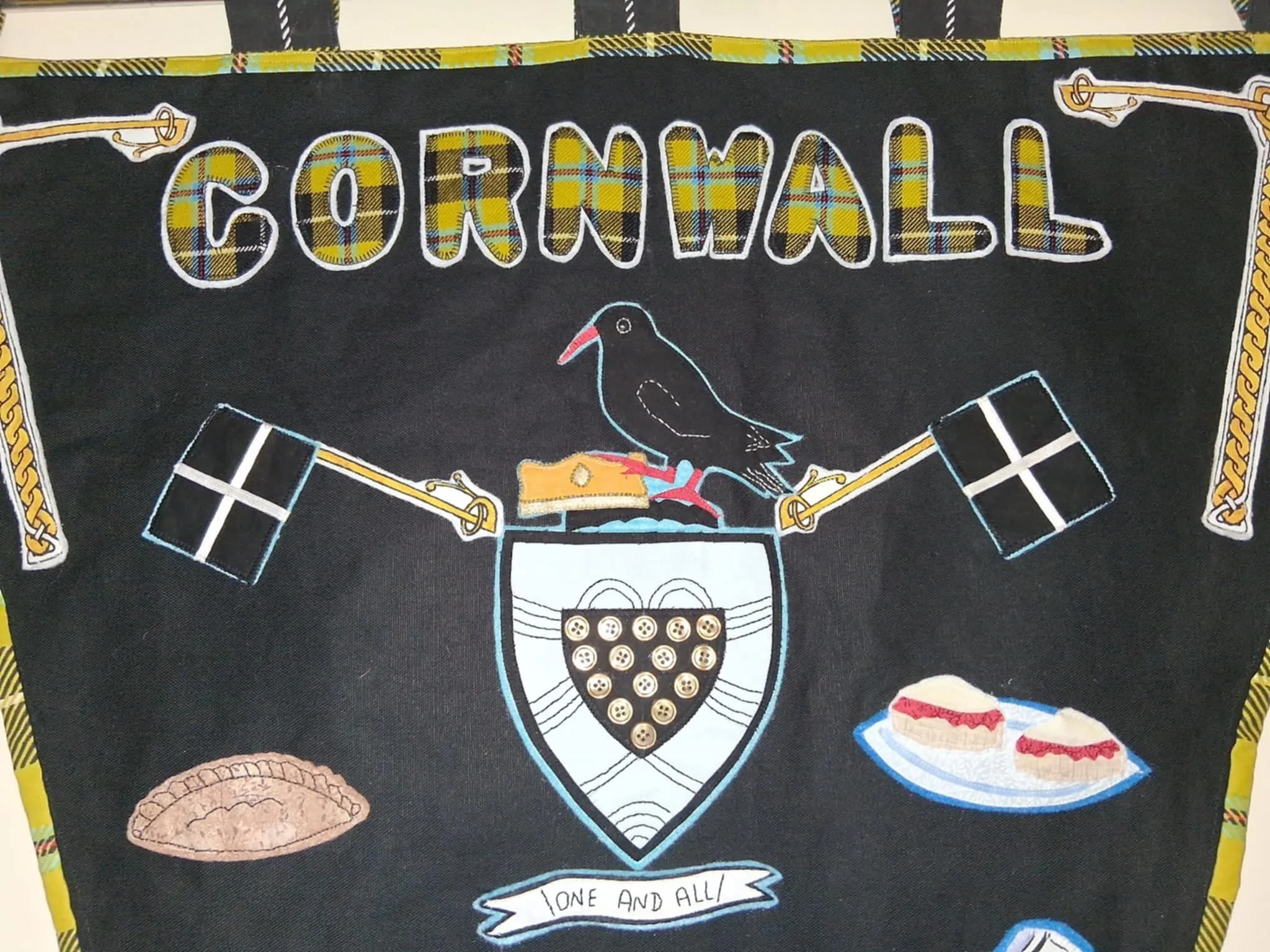 Embroidery design featuring the word 'CORNWALL' in plaid letters, a black bird with a red beak perched on a cheese wedge, a shield with buttons, a pie, and a plate of finger sandwiches, with a banner that reads 'ONE AND ALL'
