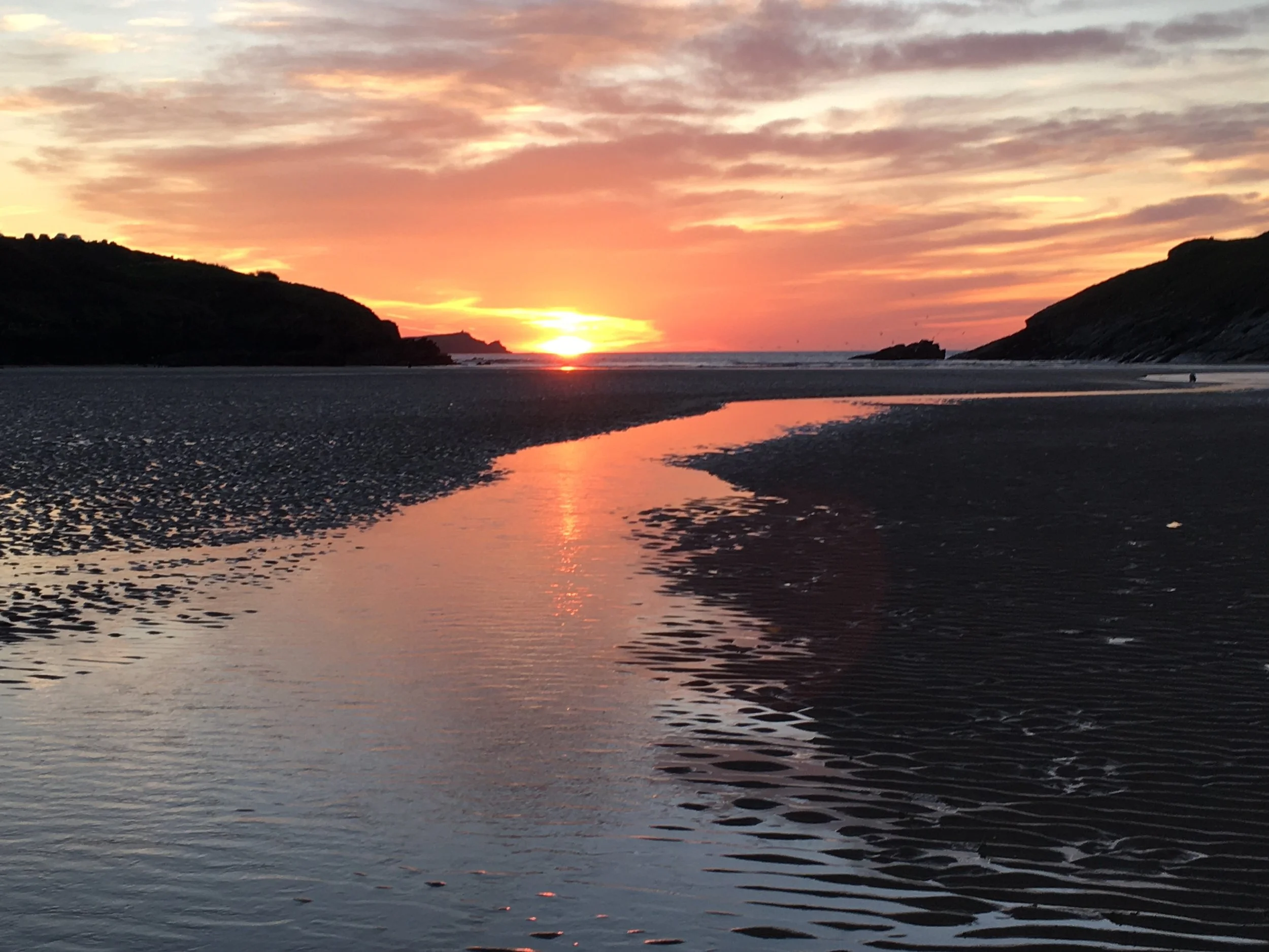 Sunset over a beach with a small stream reflecting the sky, surrounded by cliffs on both sides.