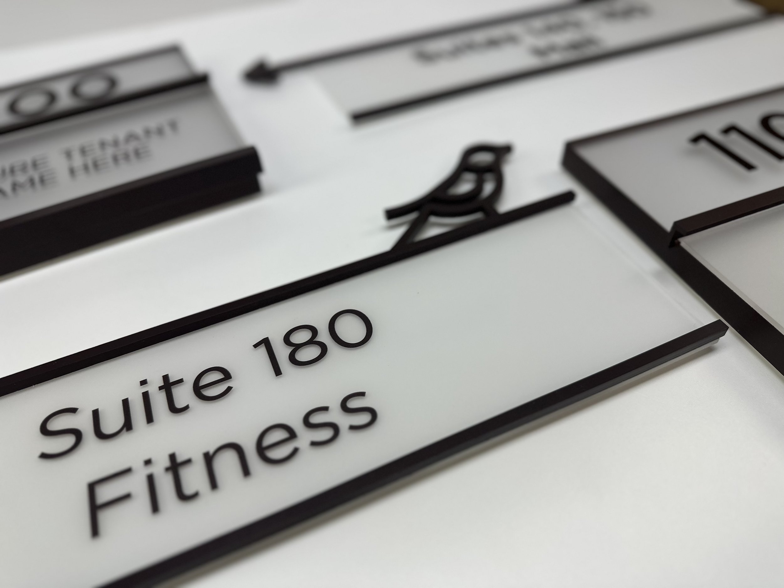 Close-up of several office suite signs on a table, featuring frosted glass with black borders and lettering. One sign reads "Suite 180 Fitness" with a small bird decoration.