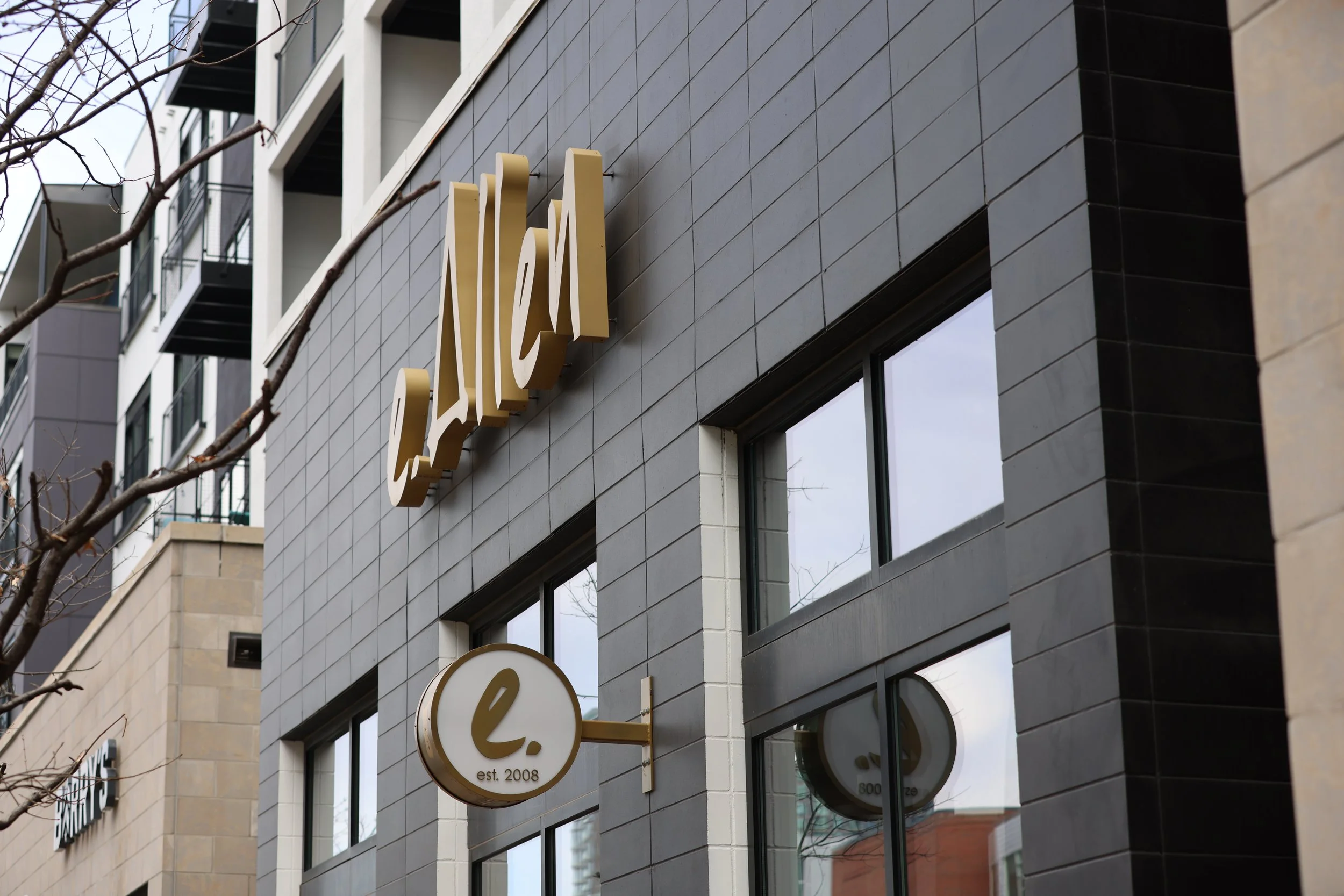 Storefront of e.Allen with a modern black tile facade, featuring gold signage and a circular logo with 'e.' established in 2008. Residential building and Barry's sign in the background.