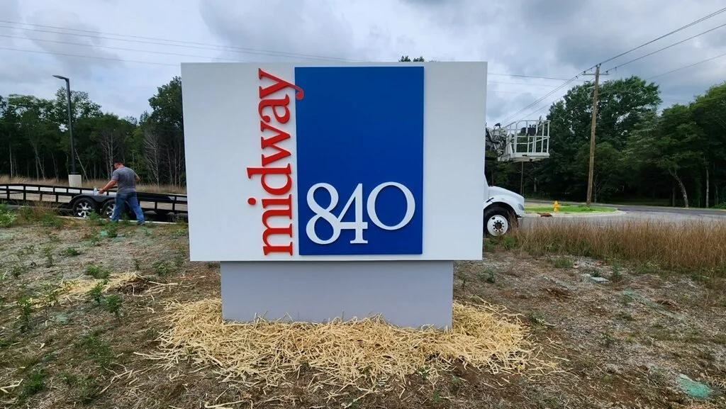 A sign with 'midway 840' on it against a backdrop of trees and cloudy sky, with a person walking and a truck in the background.