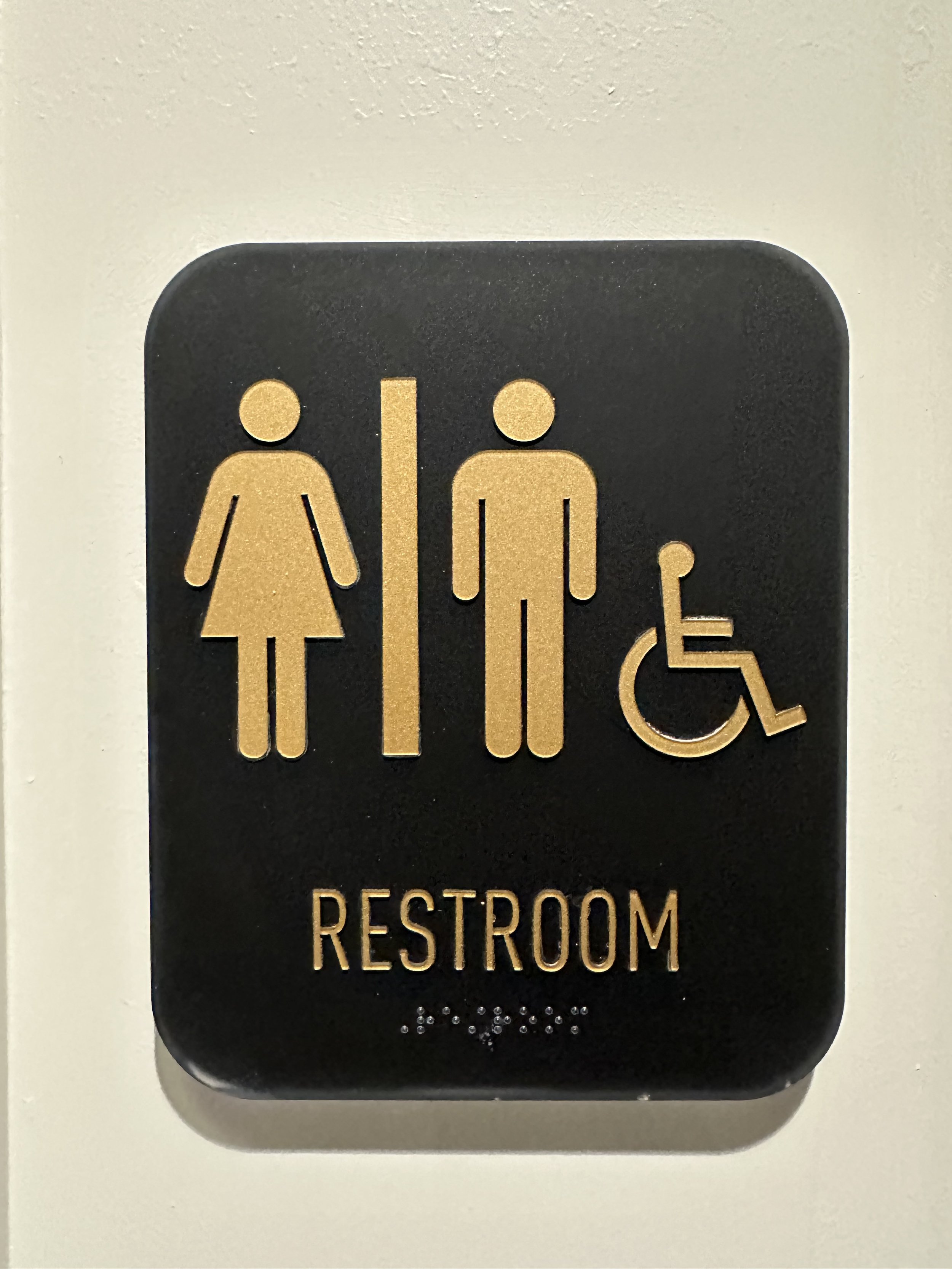 Restroom sign with symbols for men, women, and accessibility on a black background with gold-colored icons and text.