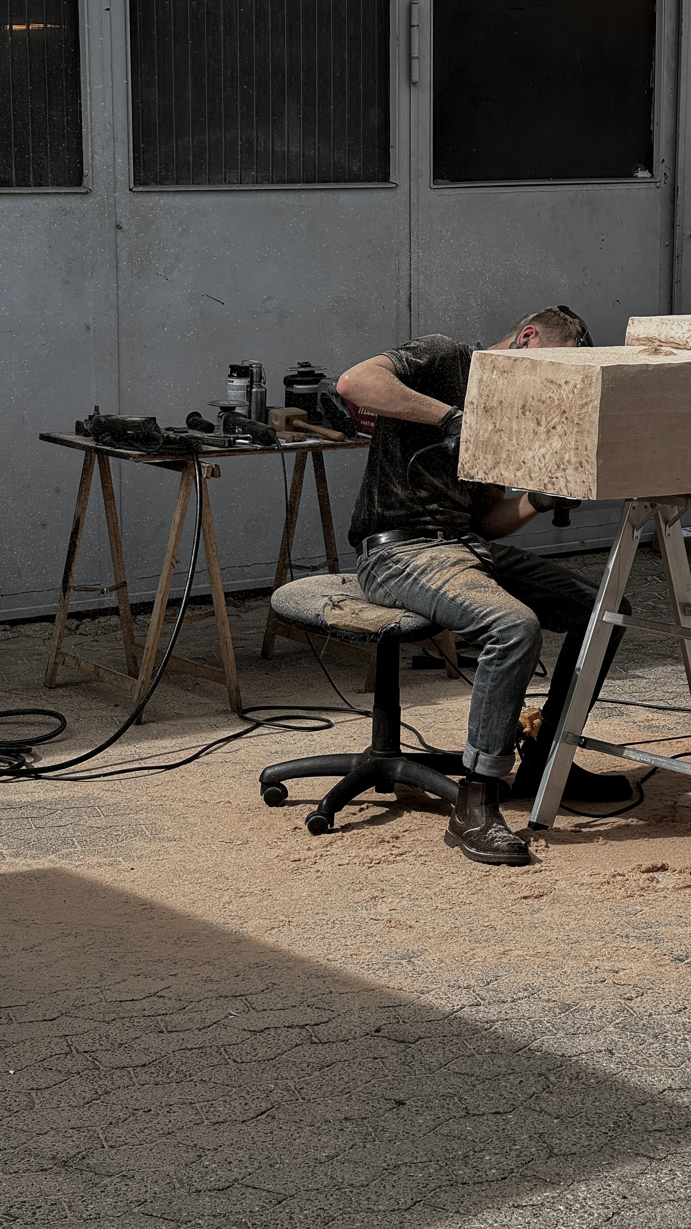A man working on a woodworking project in a workshop, sitting on a stool with tools and equipment on tables nearby, and a large piece of wood in front of him.