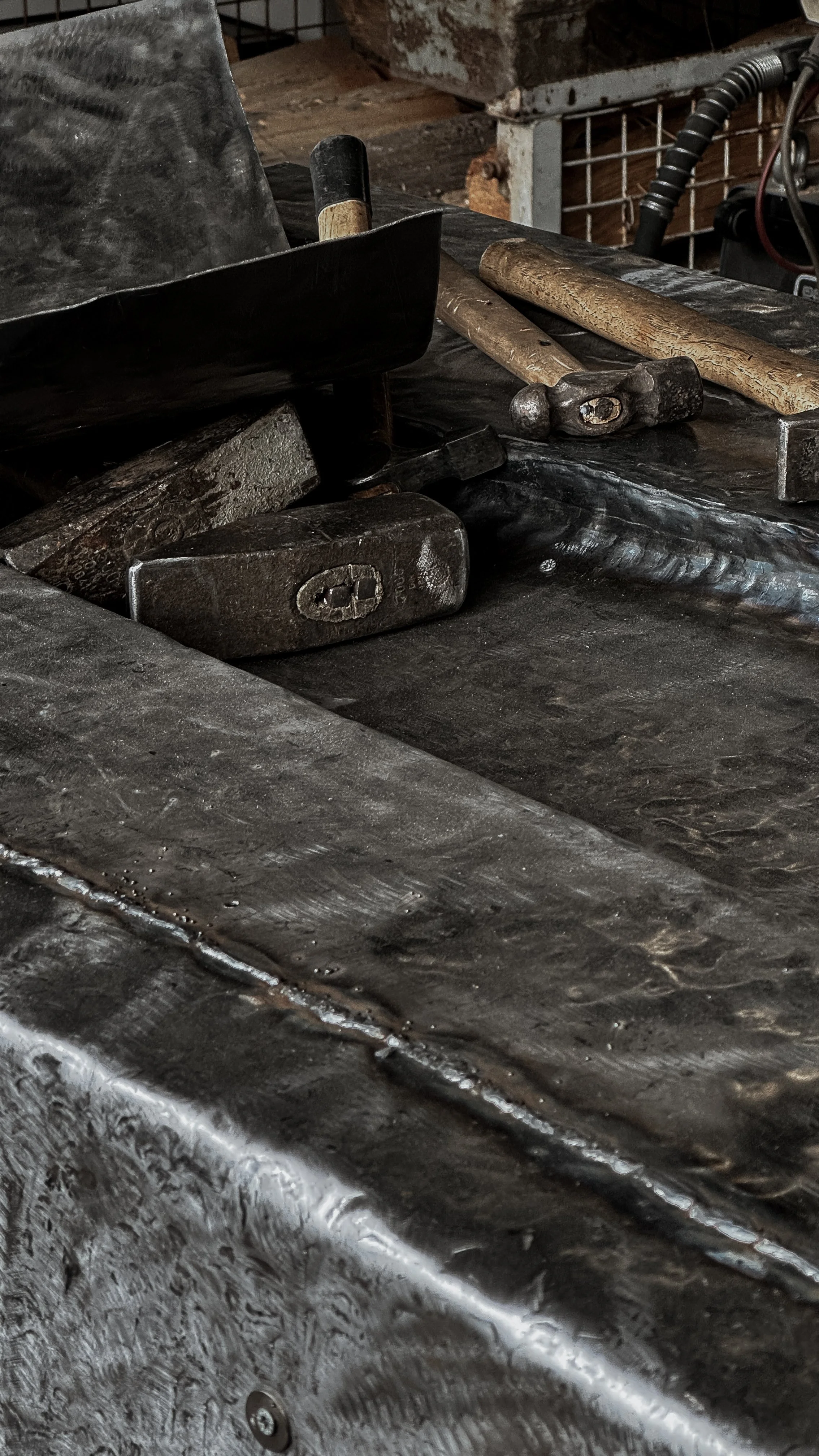 Close-up of a workbench with blacksmithing tools, including hammers and a tongs, on a dark metal surface in a forge workshop.