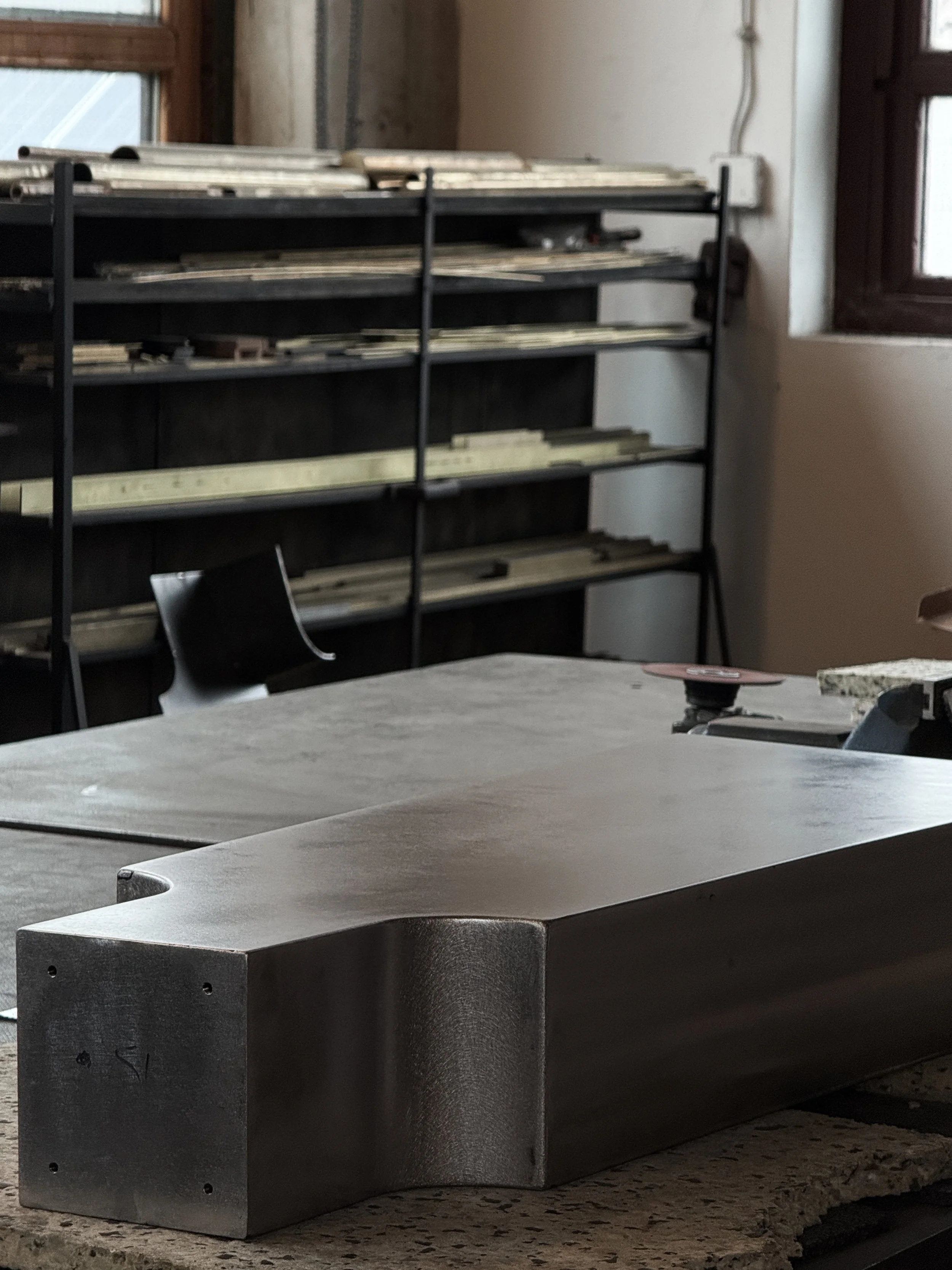 A metal piece with smooth, rounded edges on a workbench in a workshop with shelves containing metal strips and a window in the background.