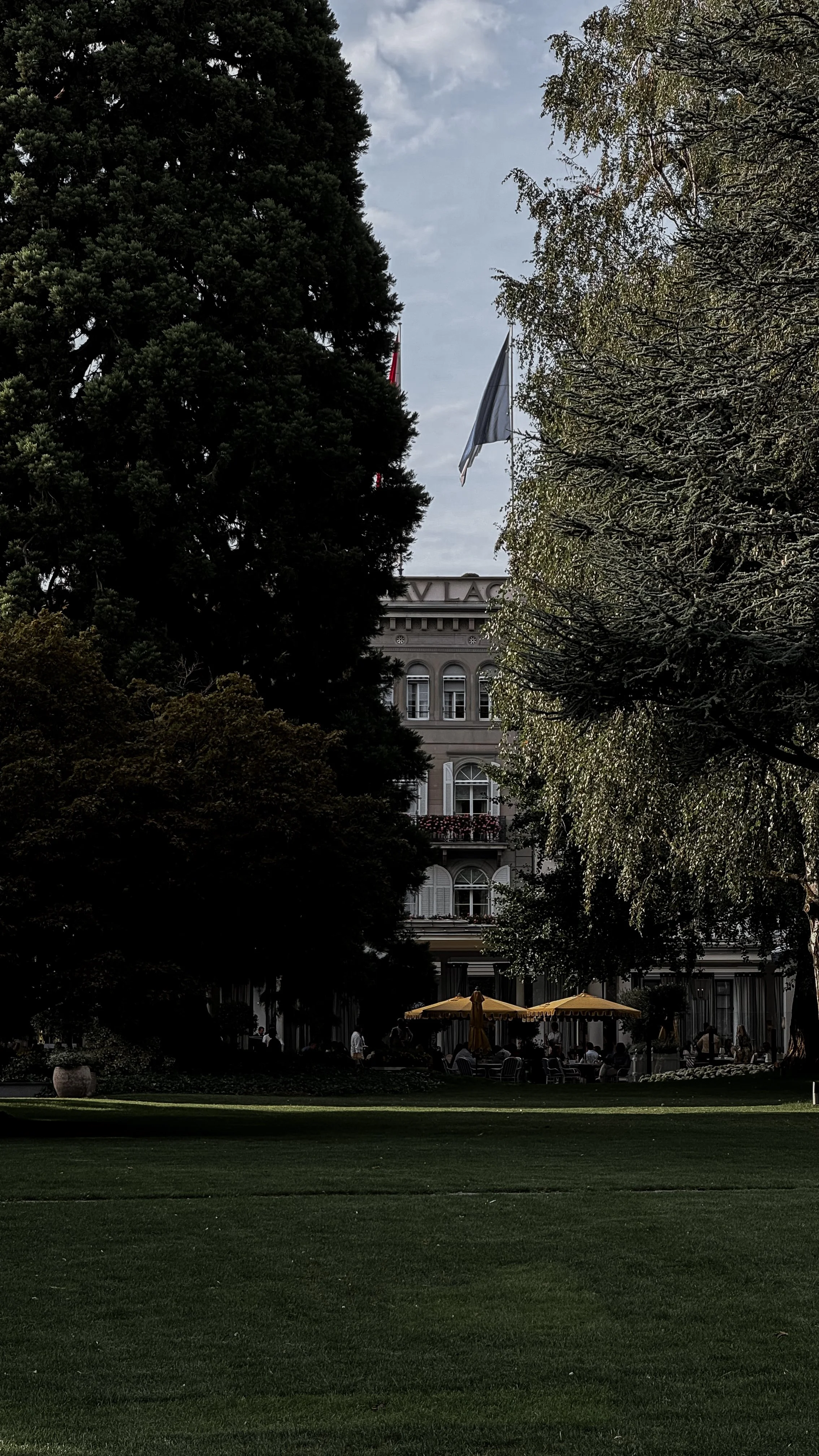 A lush green lawn in the foreground, with large trees and a historic building with a balcony and decorative windows in the background, partially obscured by trees. Outdoor umbrellas and people are visible near the building.
