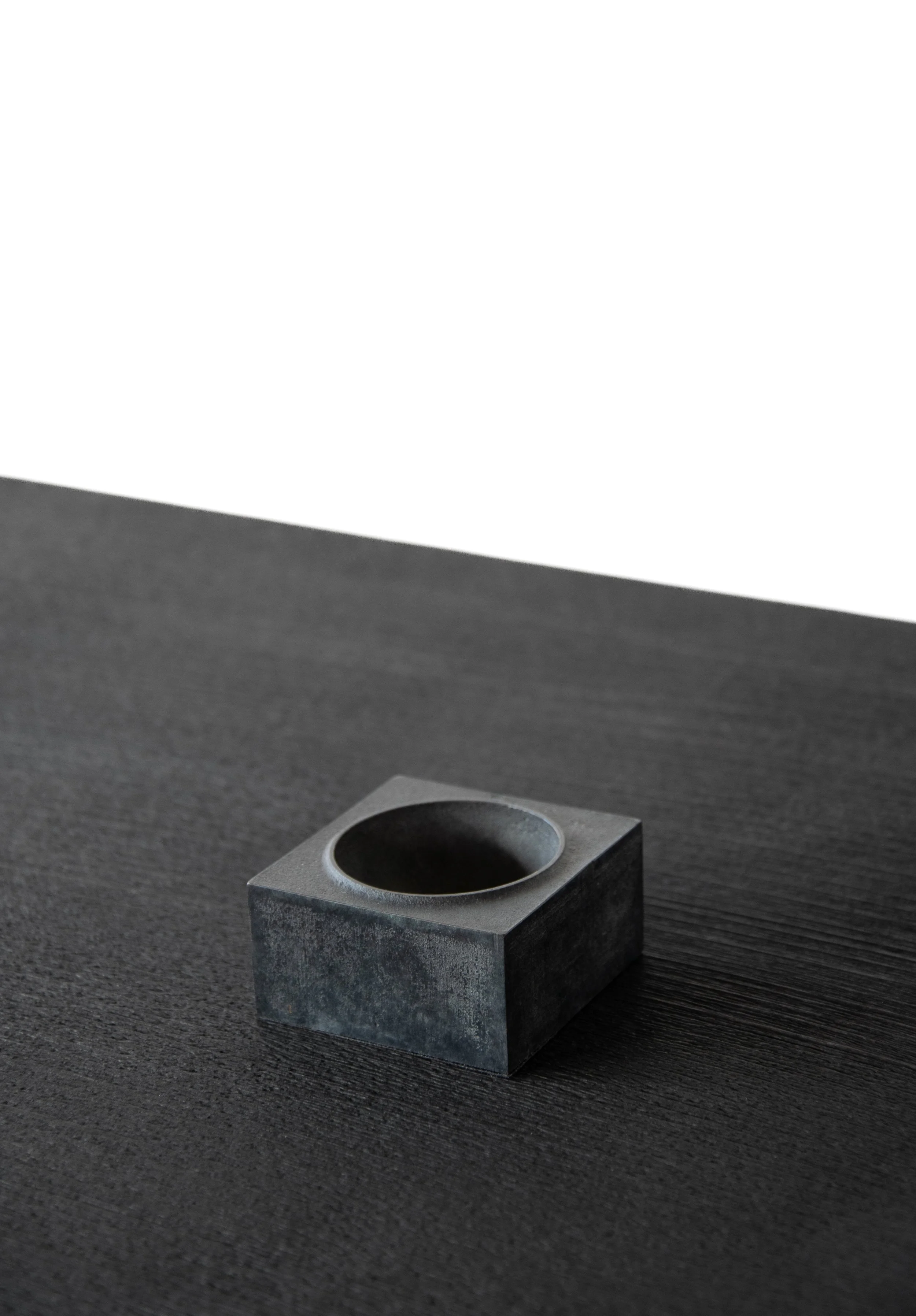 A small, matte black, square-shaped object with a round, hollowed-out center placed on a textured black surface.