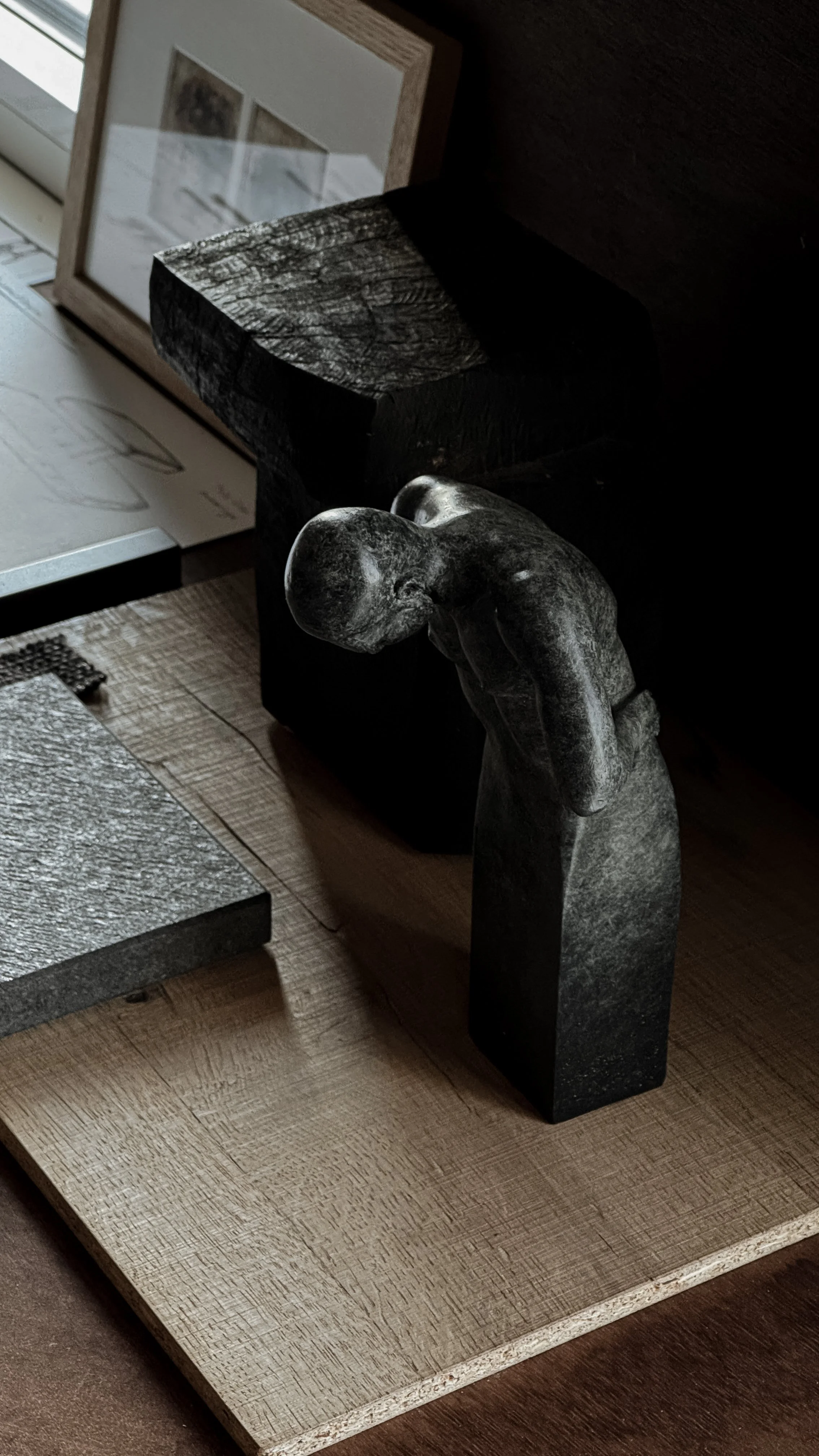 A sculpture of a bowed figure with a rounded head, placed on a wooden surface. In the background, there are framed pictures and a black block.