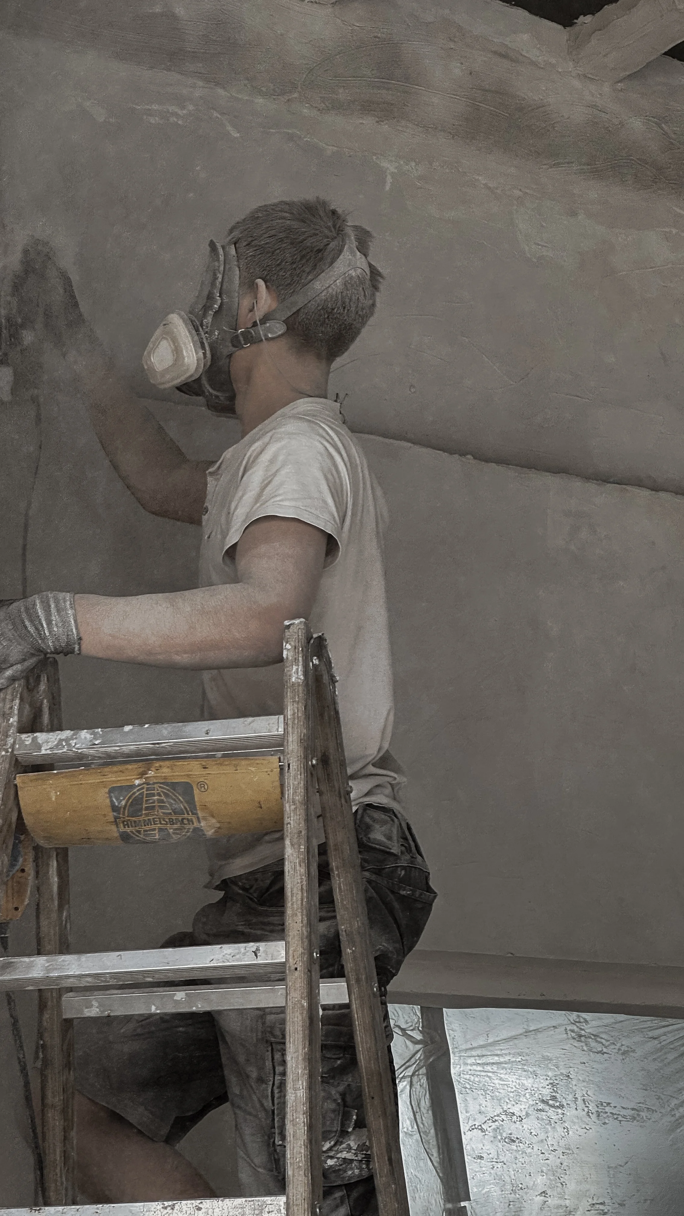 A person in protective gear, including a mask, working on a construction or restoration project on a ceiling or wall.