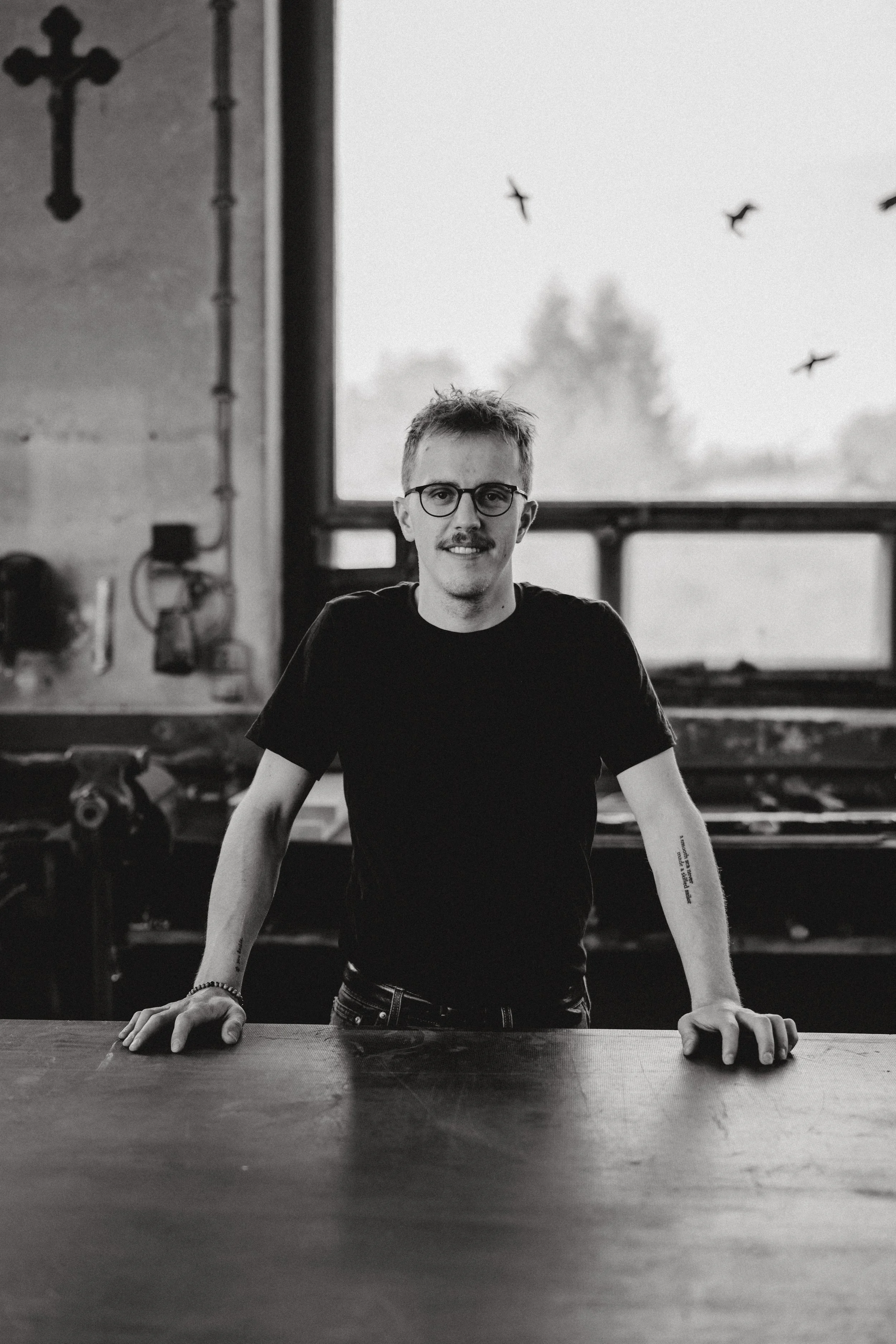 Black and white photo of a man with glasses and a mustache, standing at a table indoors, with large windows and birds flying outside in the background.