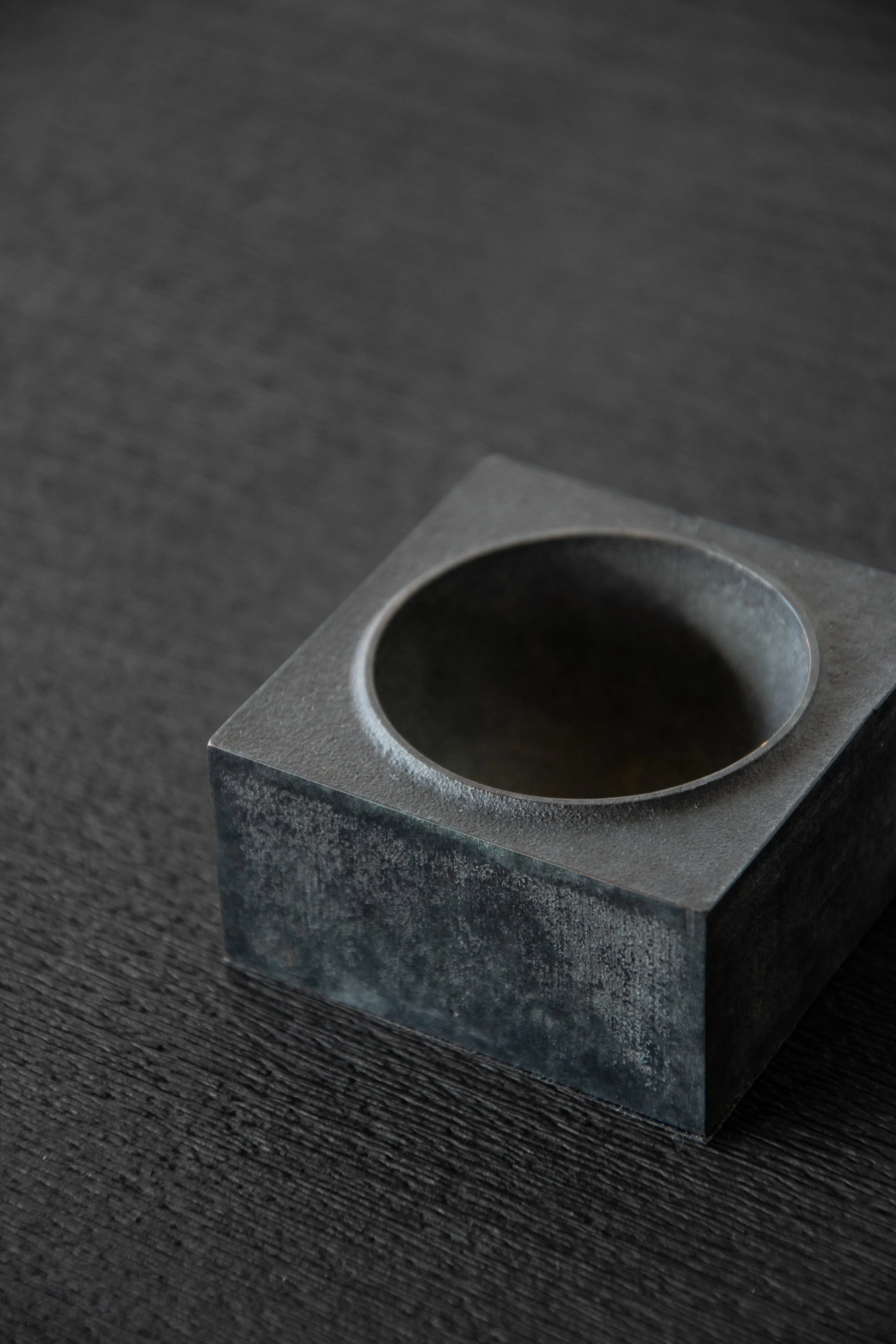 A metallic square block with a circular depression on top, placed on a dark textured surface.
