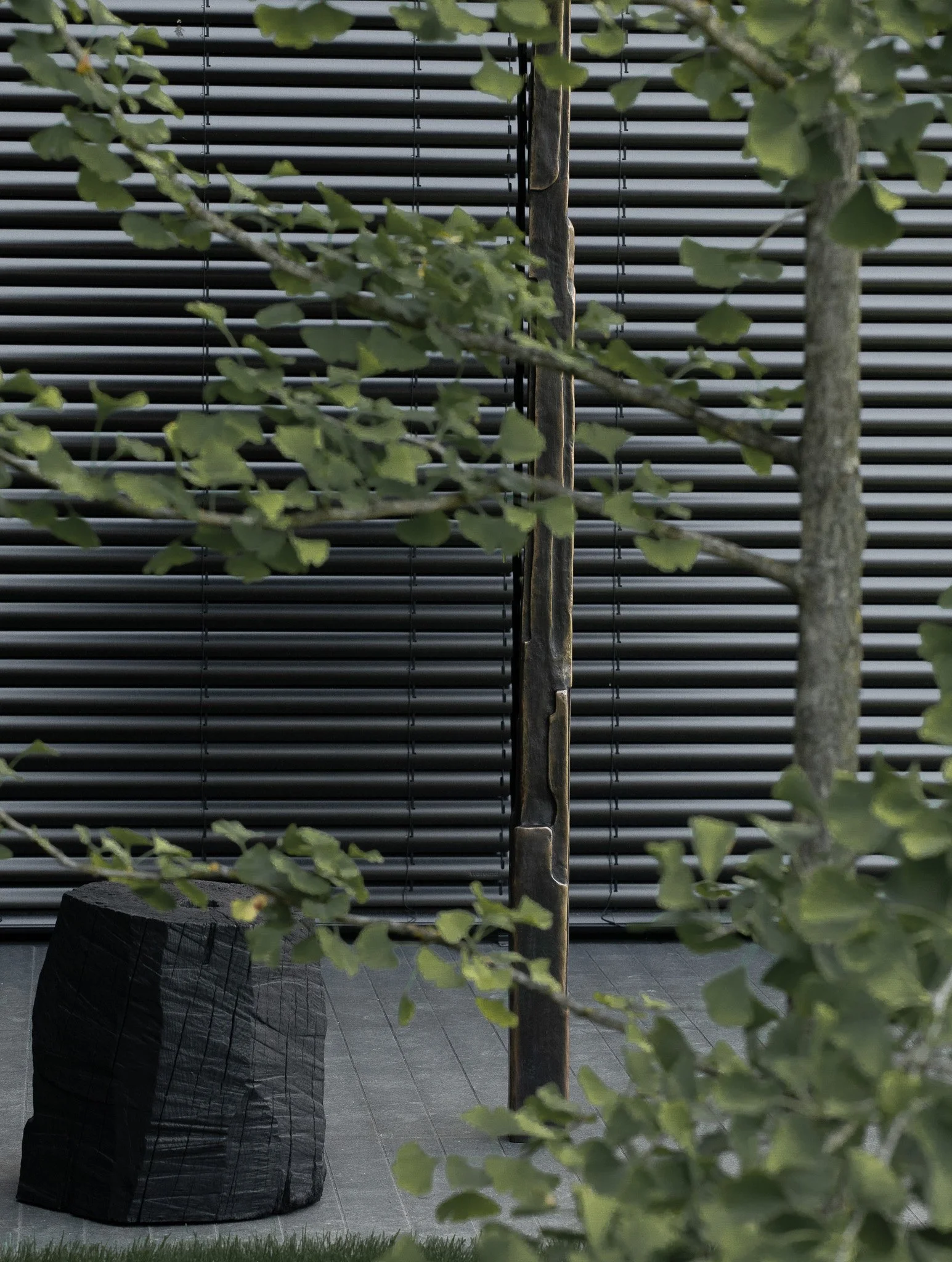 A modern outdoor space with a black stone seating block, greenery, and a metal pole, all set against a black horizontal slatted background.