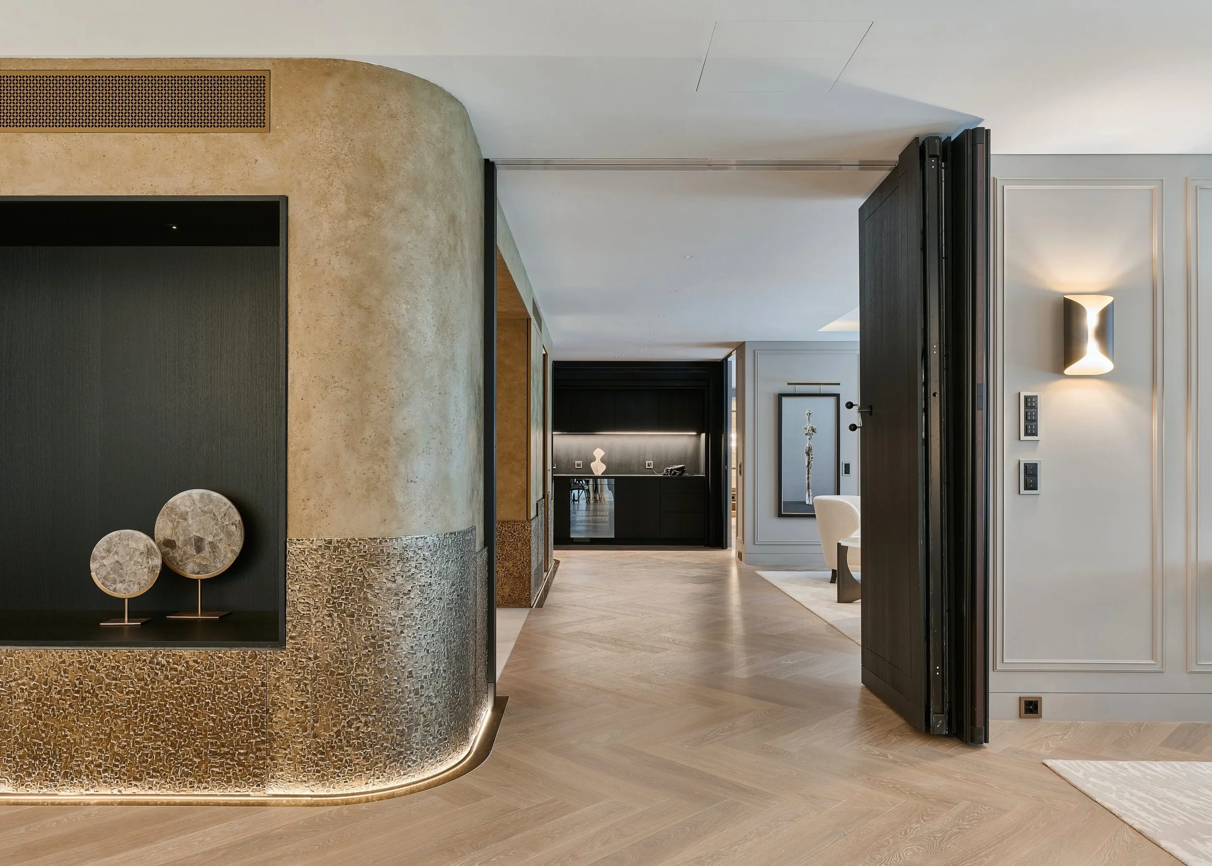 Modern interior with a curved textured wall and decorative stone circles on display, opening into a hall with light wood flooring, black panels, and white walls with accent lighting.