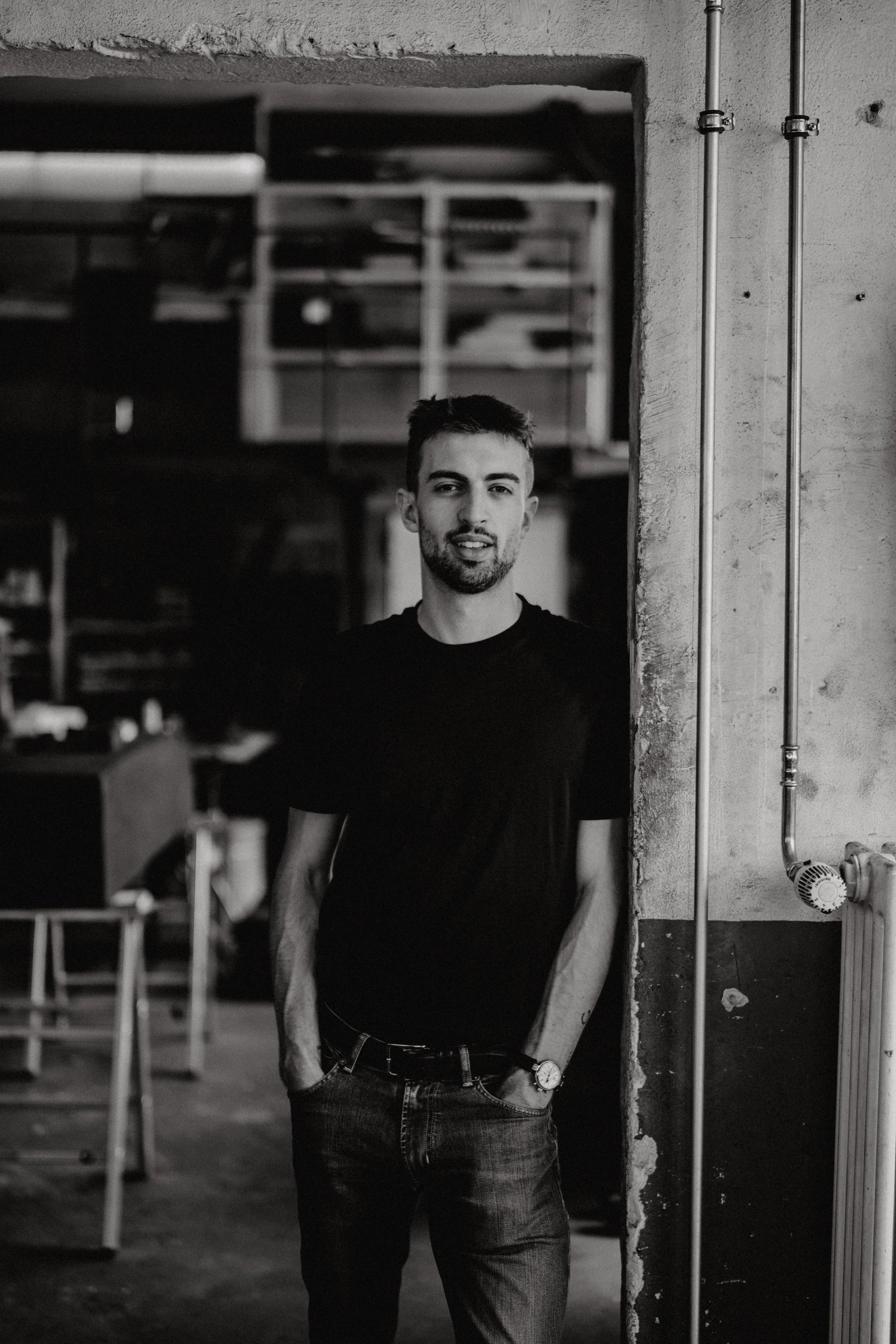 A young man with short hair and a beard standing in a warehouse or industrial space, leaning against a concrete wall with pipes, wearing a black t-shirt and jeans, with a watch on his left wrist.