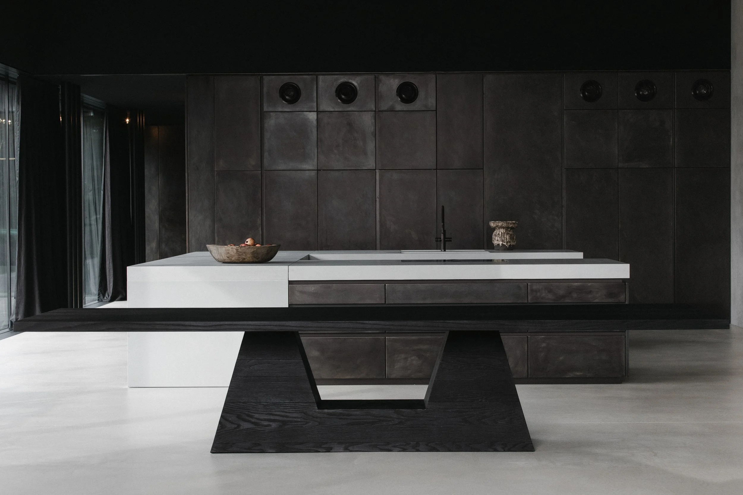 Modern kitchen with dark cabinets and a white island, featuring a black table in the foreground, a bowl of fruit, and decorative objects.