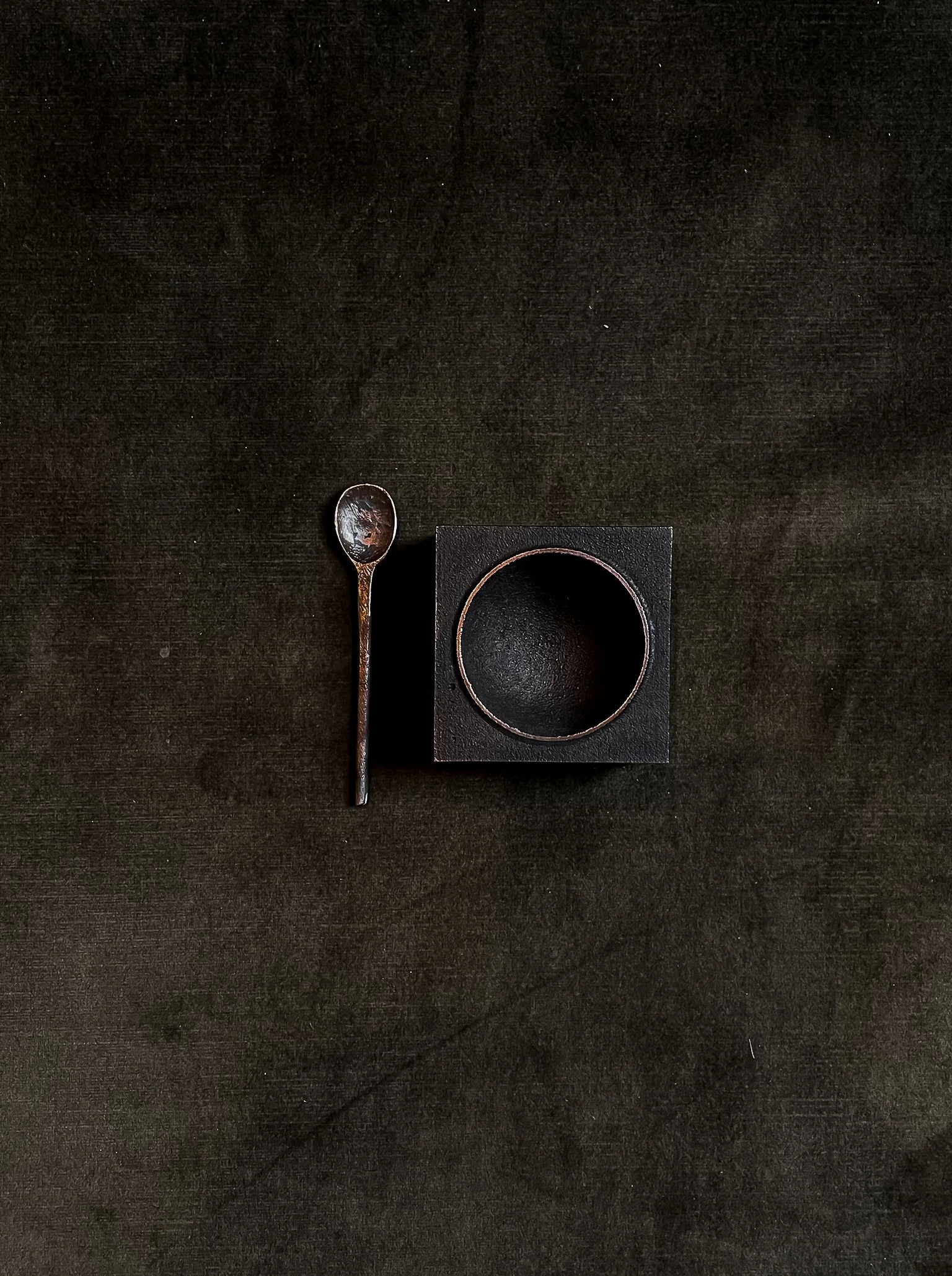 A black square container with a circular opening, a small metallic spoon to its left, all placed on a dark textured surface.