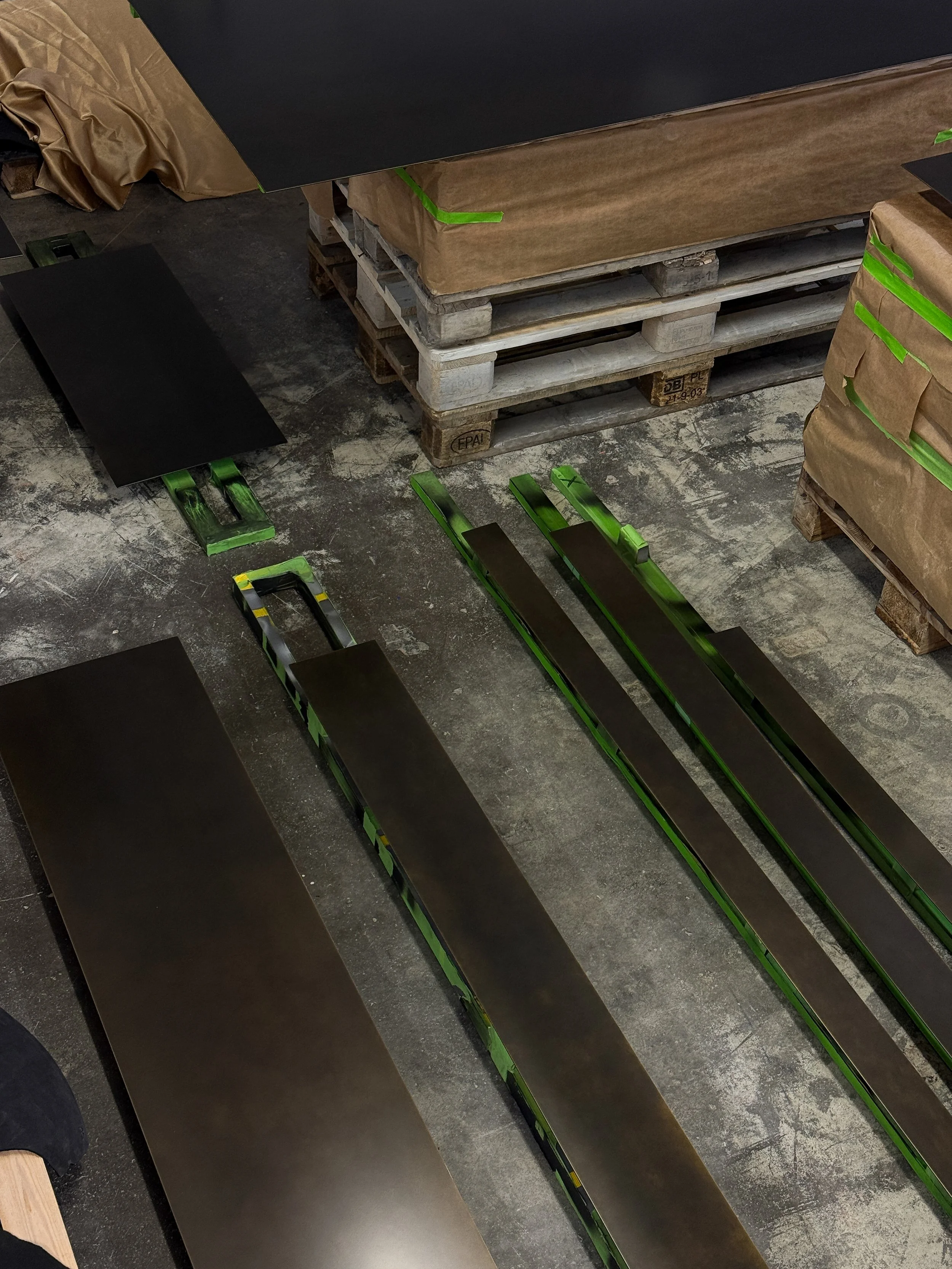 Black planks on green and black saw horses, stacks of wrapped wooden pallets, and dusty concrete floor.