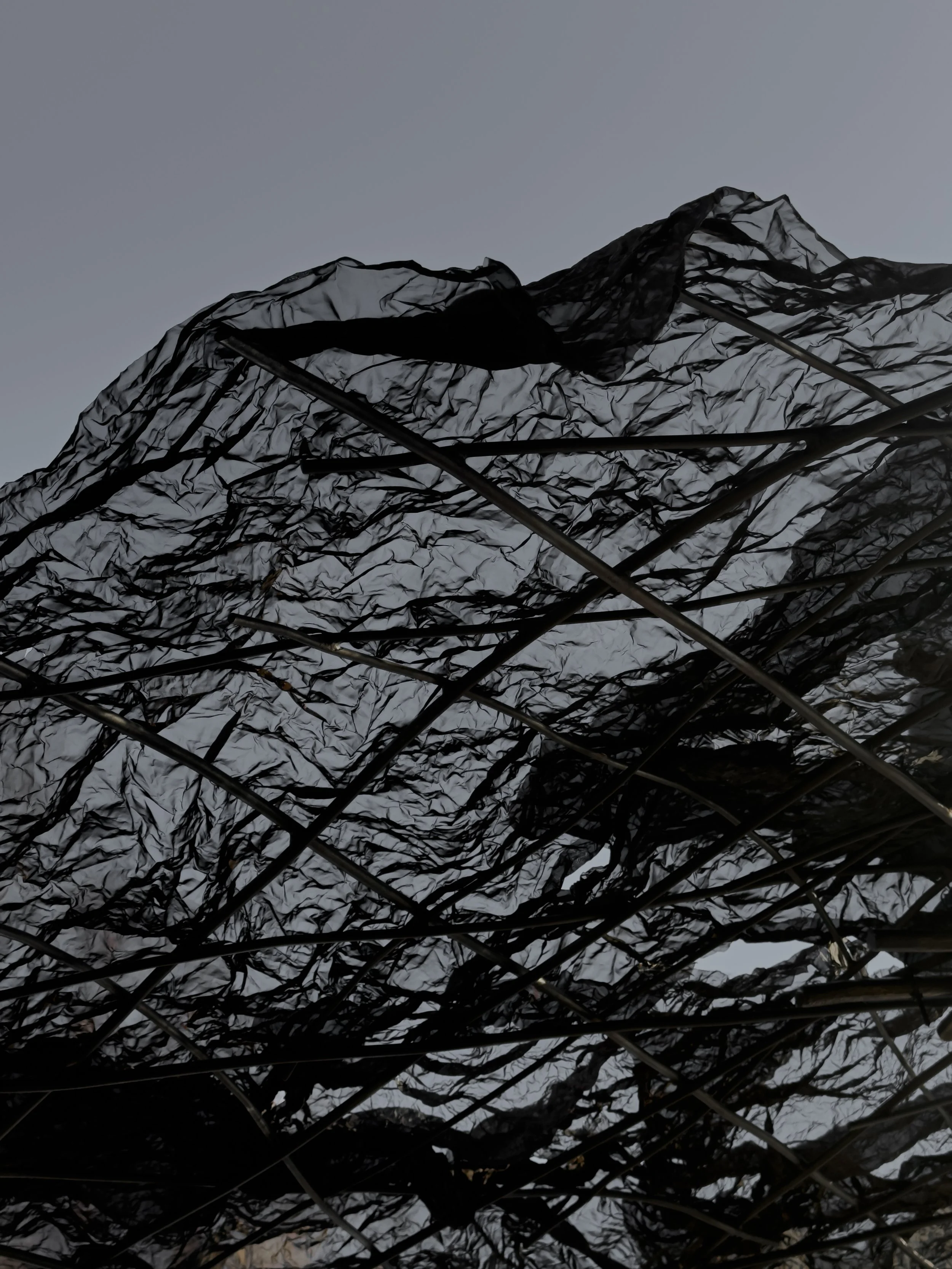 Close-up of a crumpled black plastic sheet or tarp with metal rods underneath, against a gray sky.