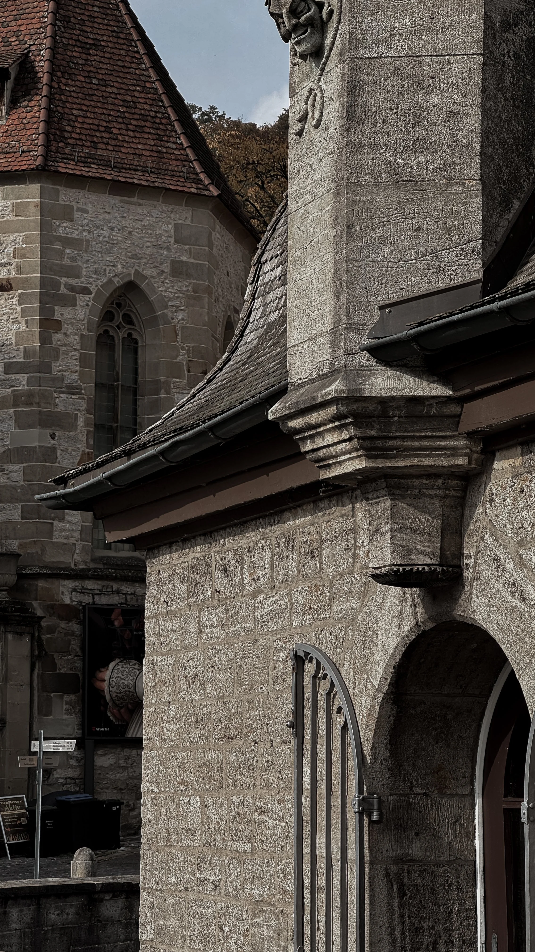 Close-up of the stone wall and roof of a historic building with a decorative stone ledge and a metal gate, featuring a large Gothic-style window in the background.