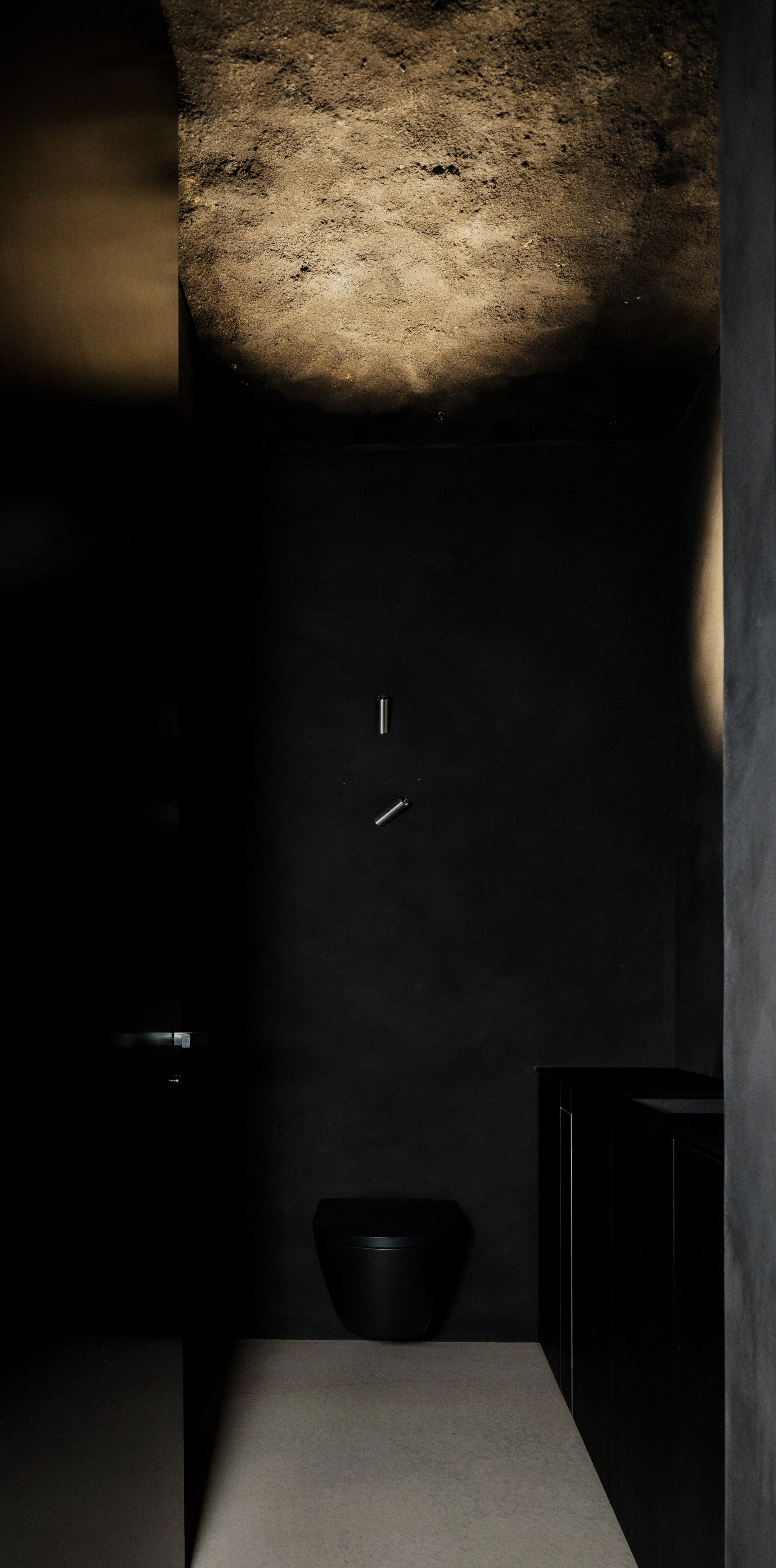 Minimalist bathroom with a black toilet, black cabinetry, and a textured ceiling with a dark wall behind. Small metallic objects are mounted on the wall.