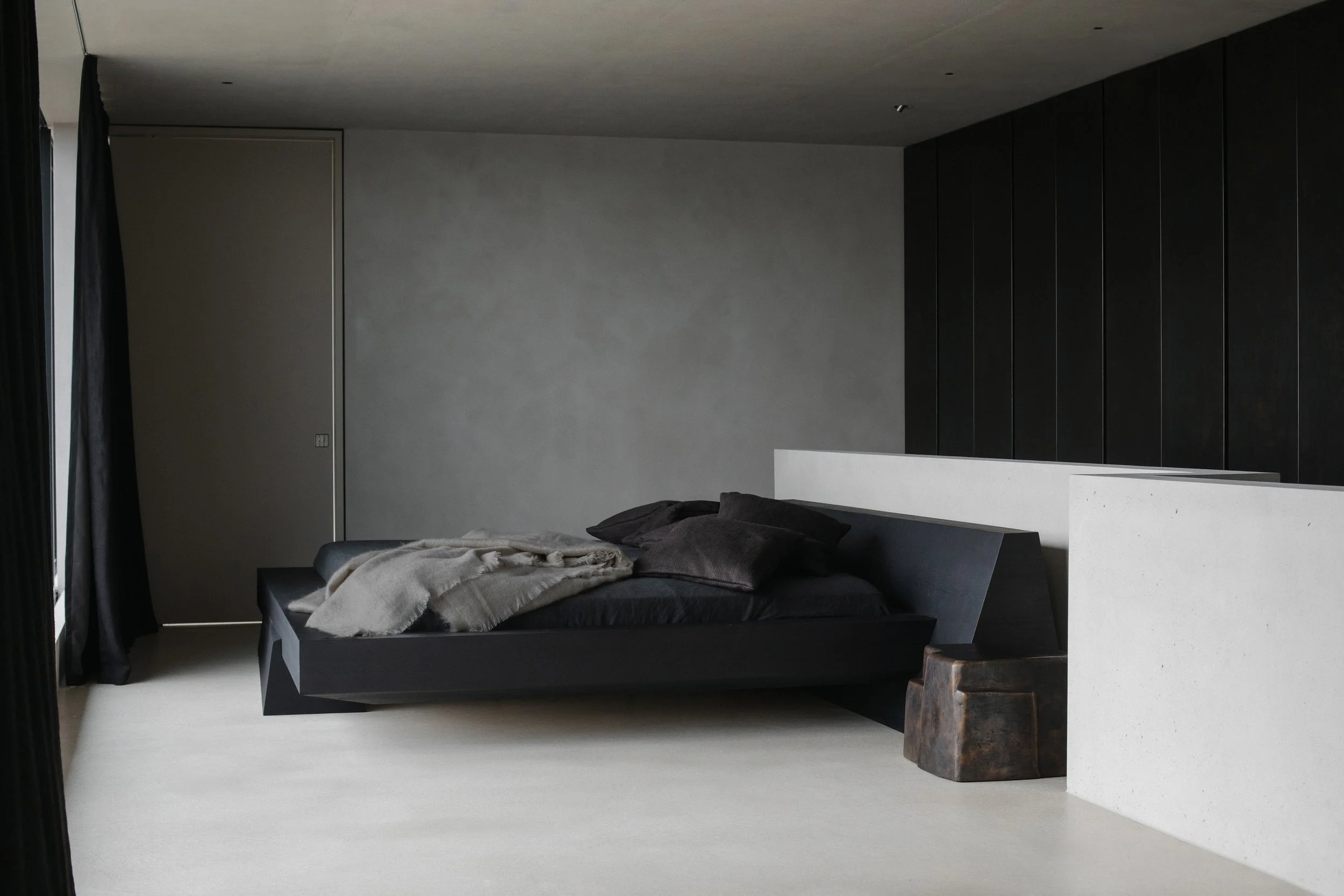 Modern bedroom with black bed, dark pillows, beige blanket, gray walls, black closet, and a small brown side table.