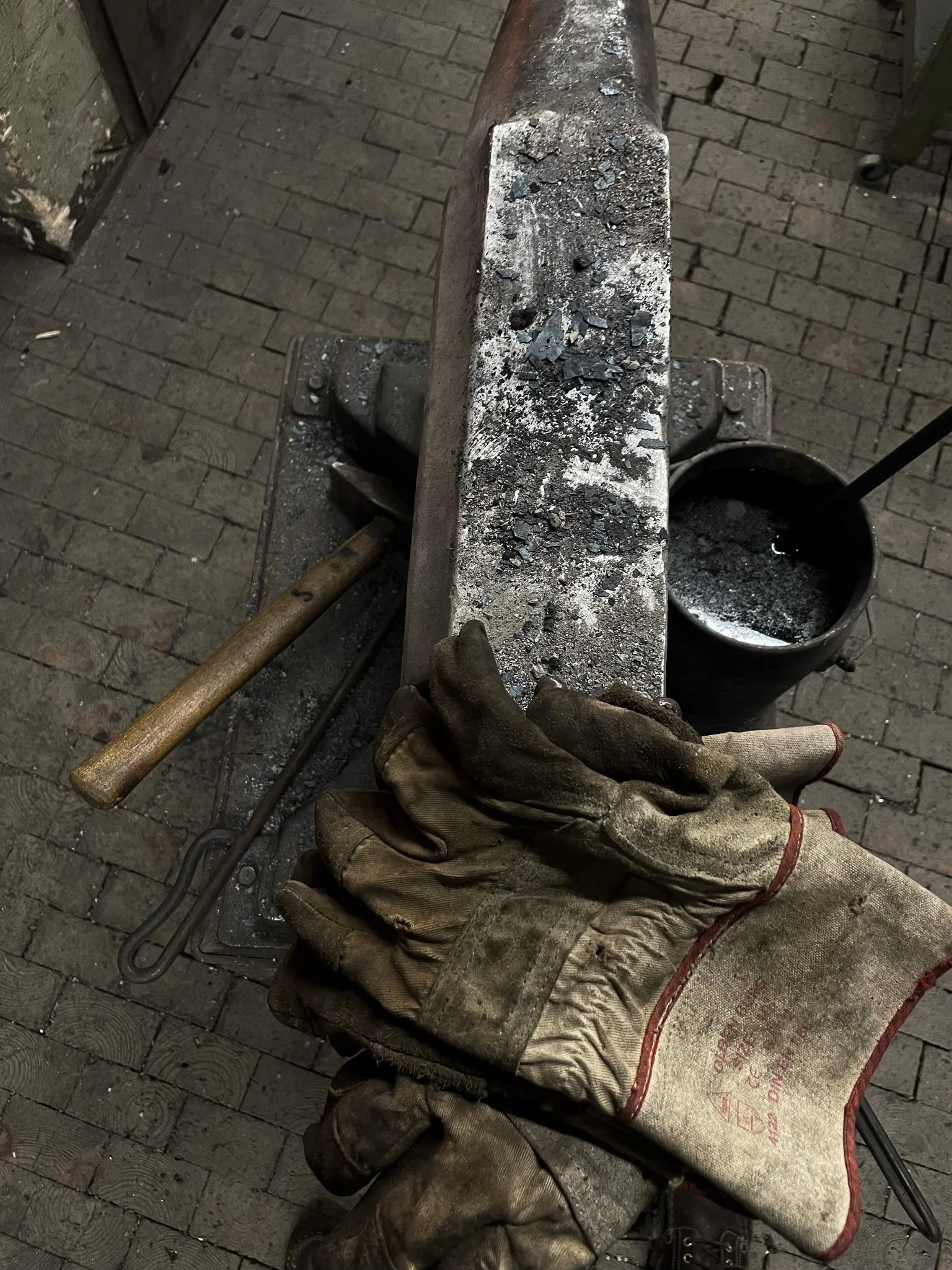 Blacksmithing work area with a metal anvil, a pair of worn leather gloves, and a container of black molten metal or slag.