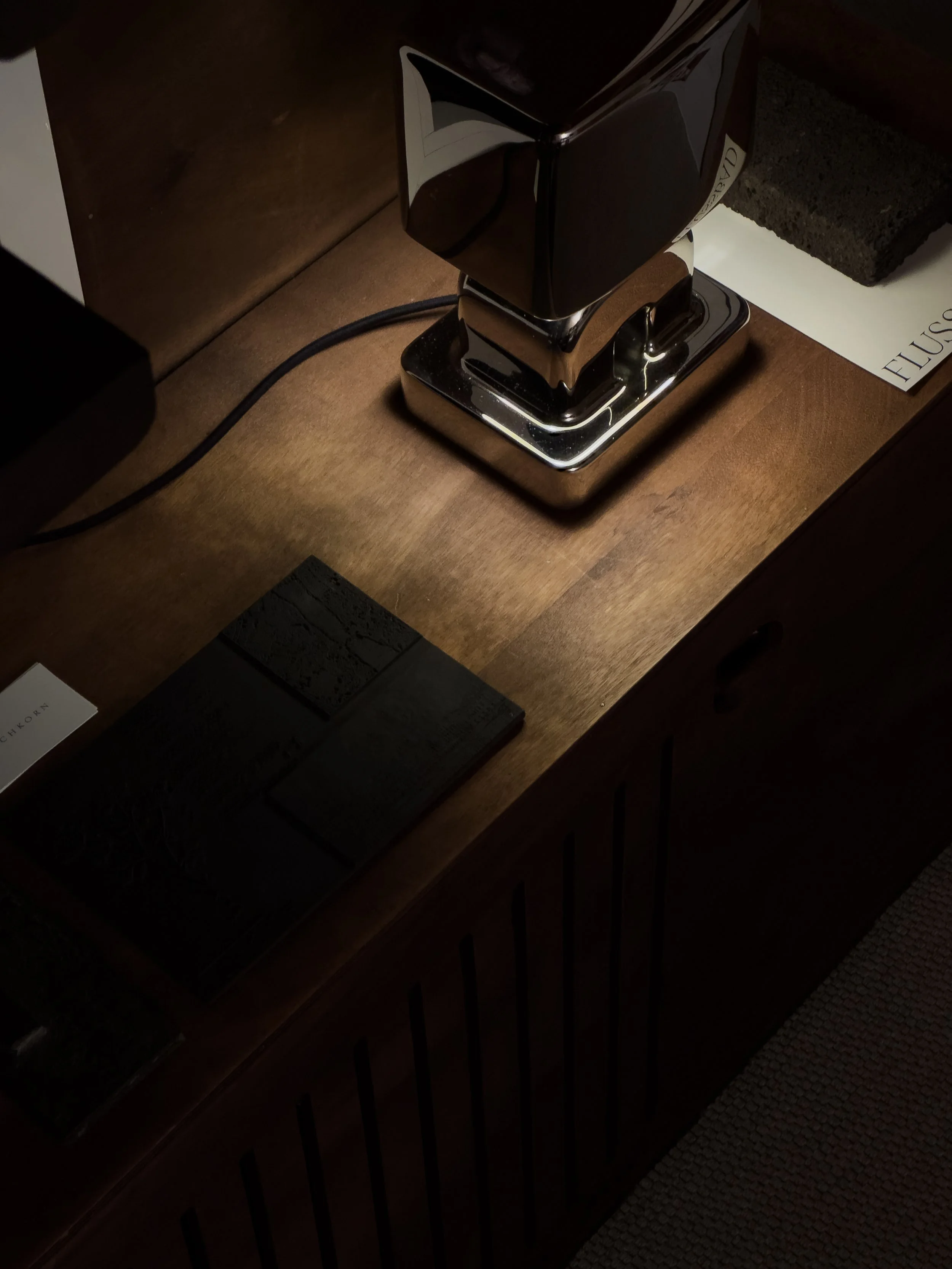 A wooden surface with a shiny, chrome table lamp, a black rectangular object, and some books or magazines nearby.