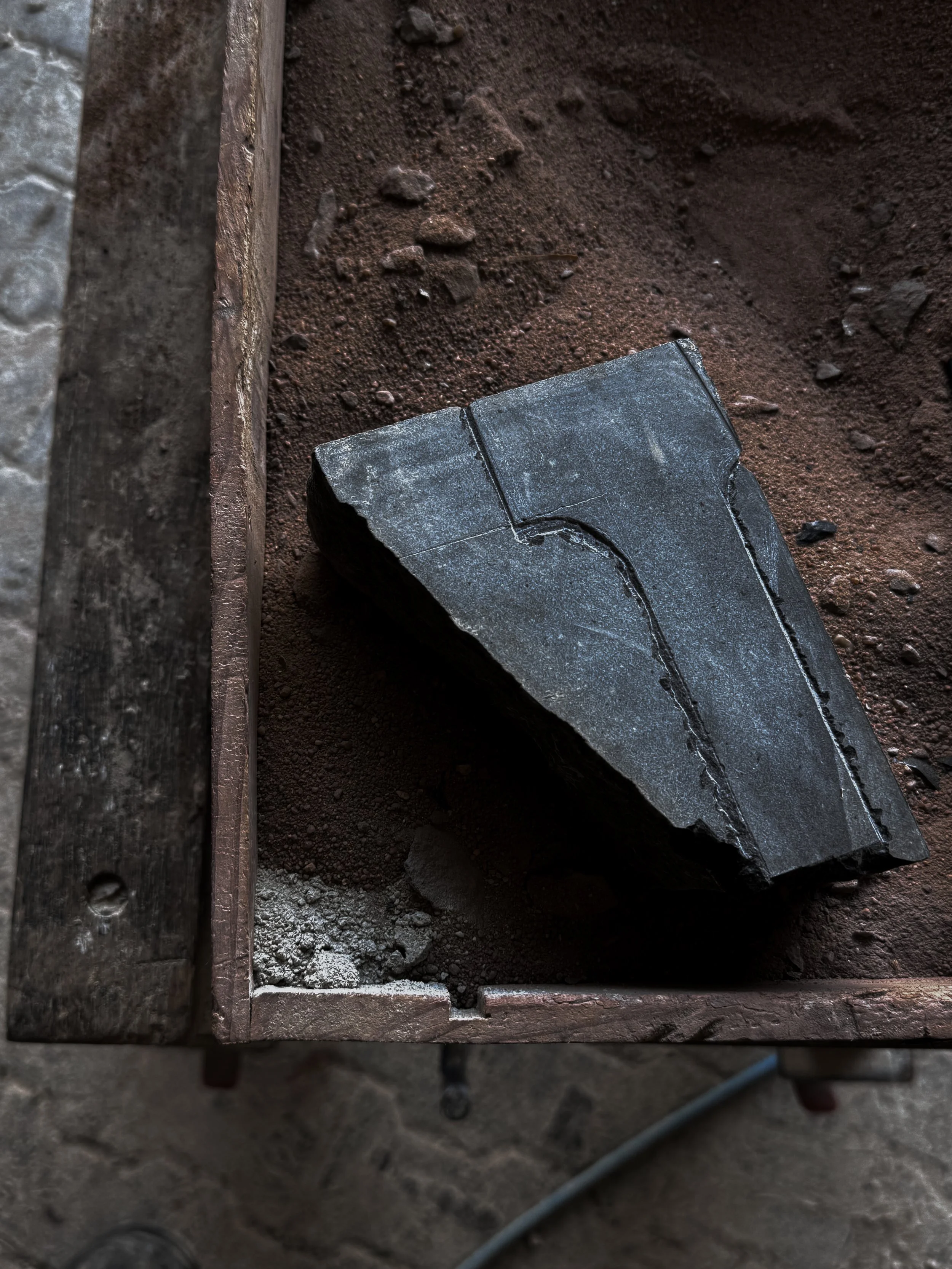 A rough, irregularly shaped piece of black stone with white veins lies on a bed of reddish-brown dirt inside a wooden frame.