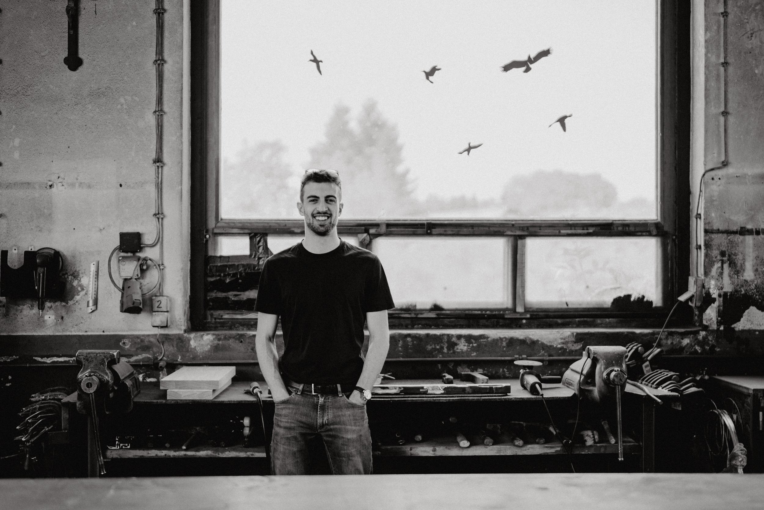 A smiling man stands in a workshop with tools on a workbench, large window behind him with birds flying outside, black and white photo.