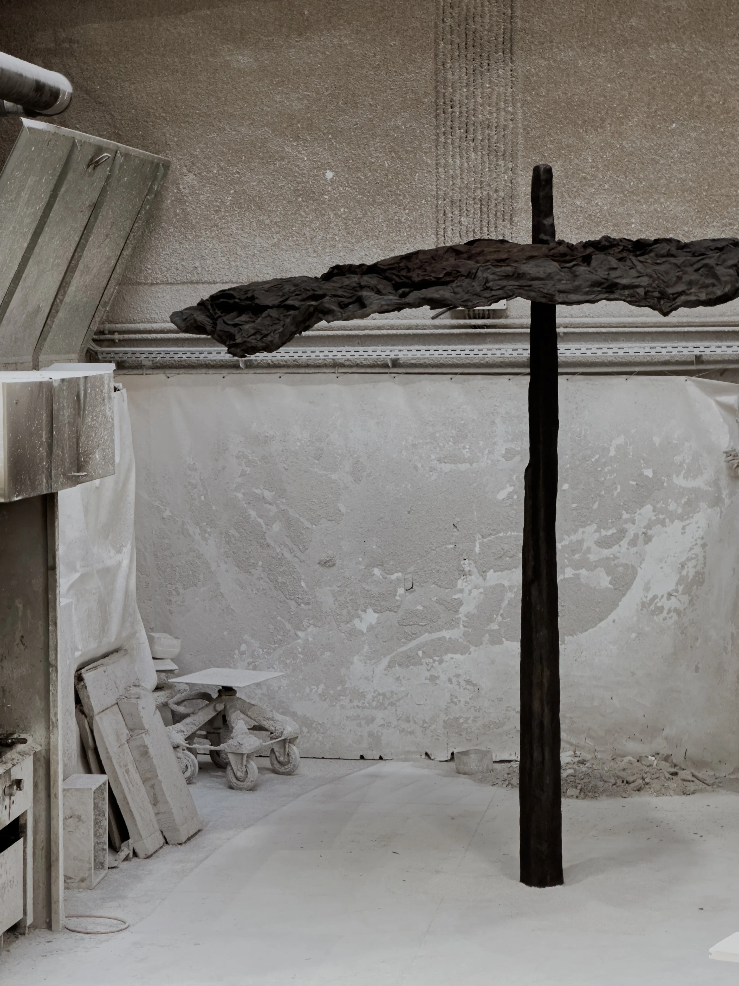 A black-painted cross with a textured horizontal arm, standing on a dusty, unfinished floor against a rough, uneven wall in a workshop or art studio.