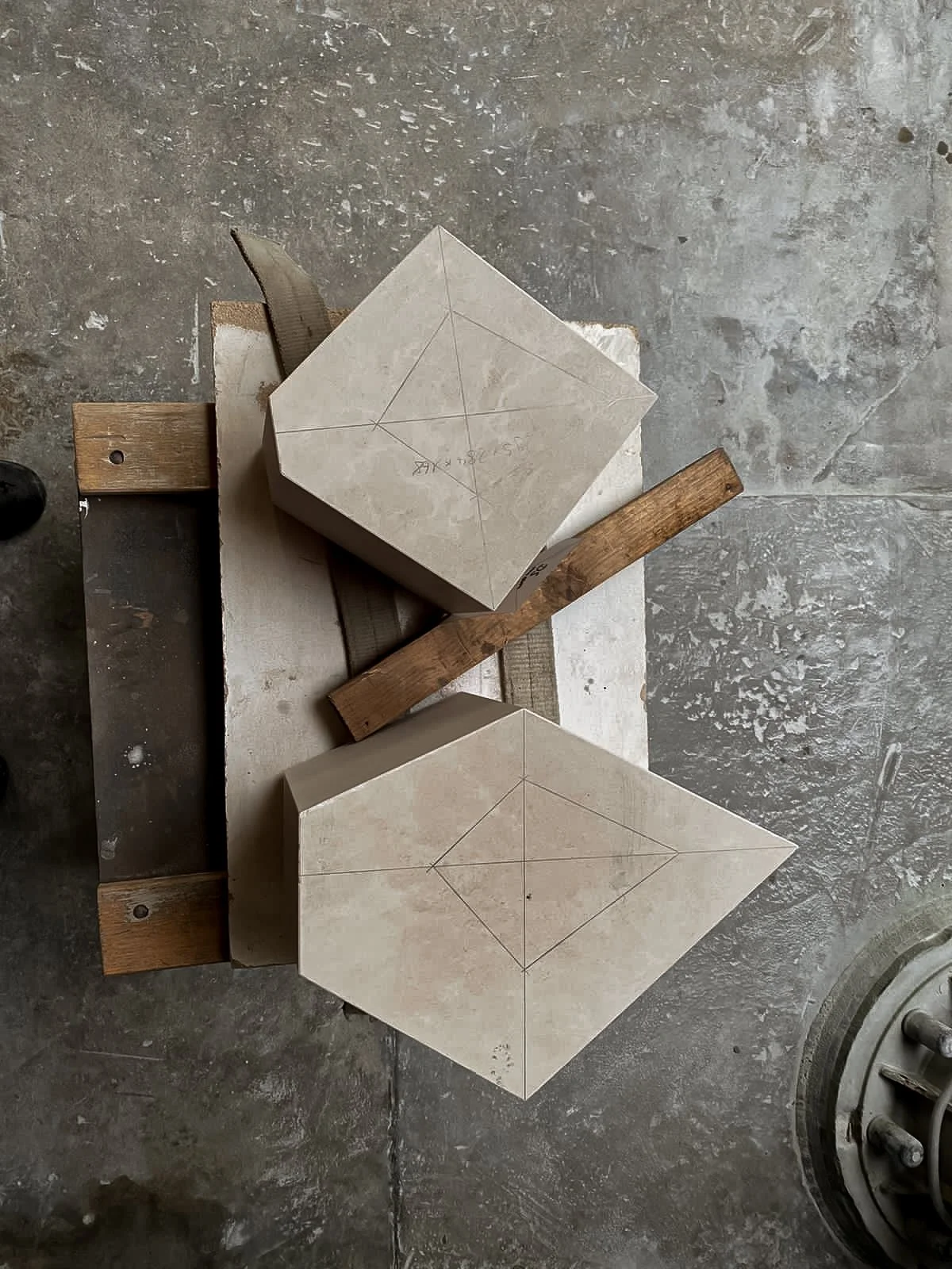 Two stone blocks with pencil sketches for cutting, placed on a small wooden and metal structure with a concrete floor in the background.