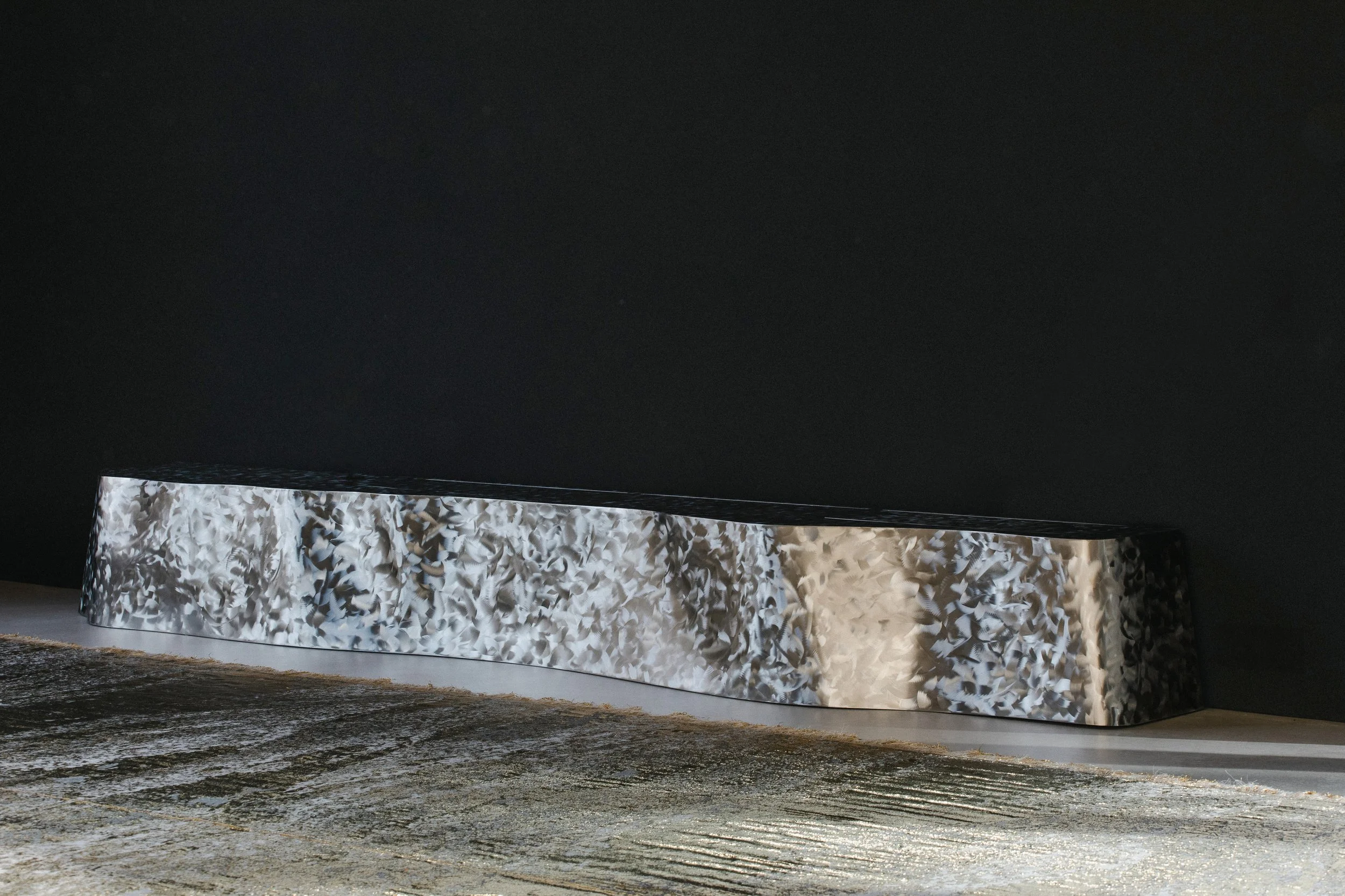 Modern metallic sculpture with textured, silver, patterned surface against a black background.