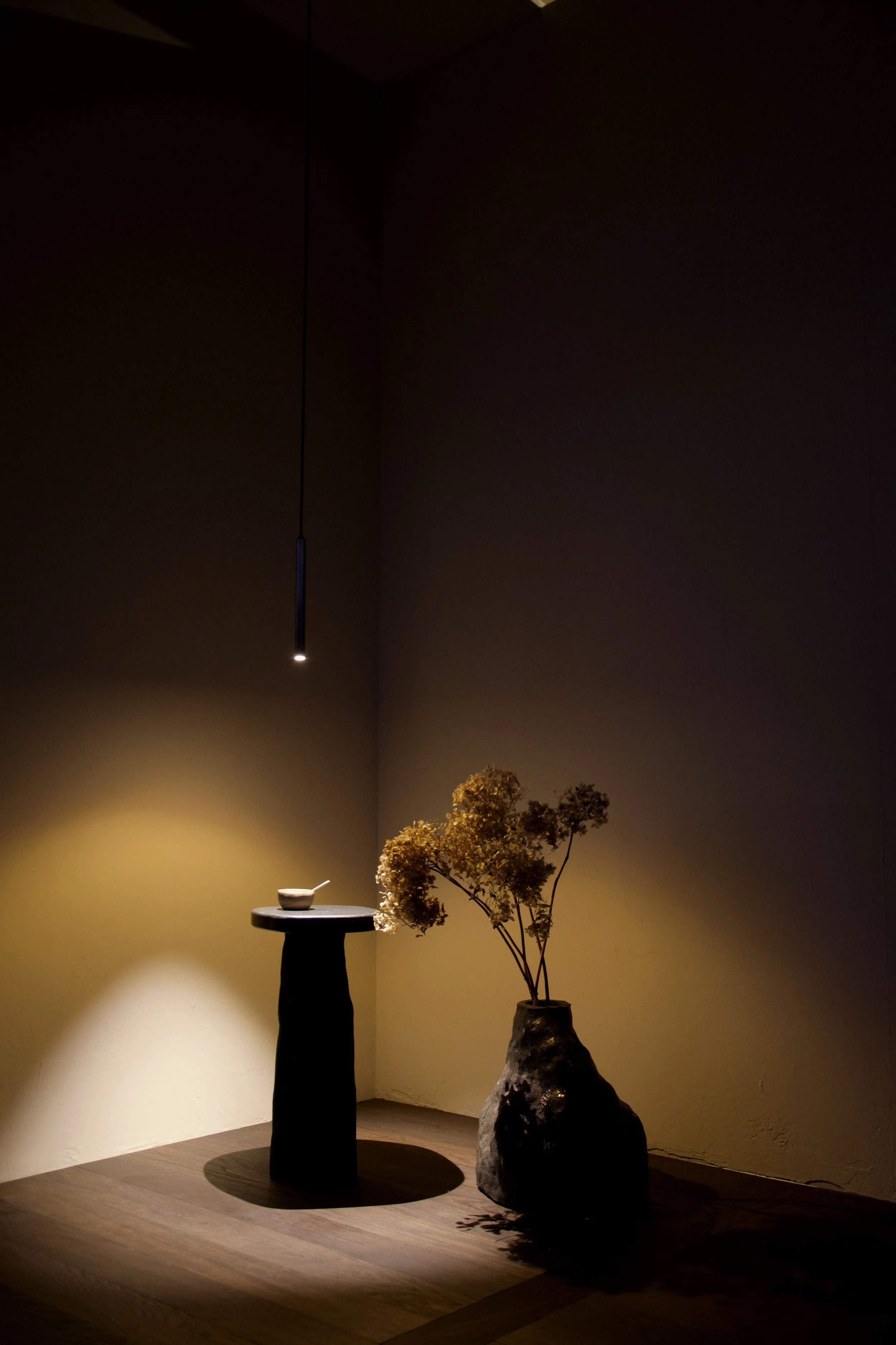 A dimly lit room with a black pedestal table holding a bowl and spoon, a large rustic vase with dried flowers, and a hanging pendant light.