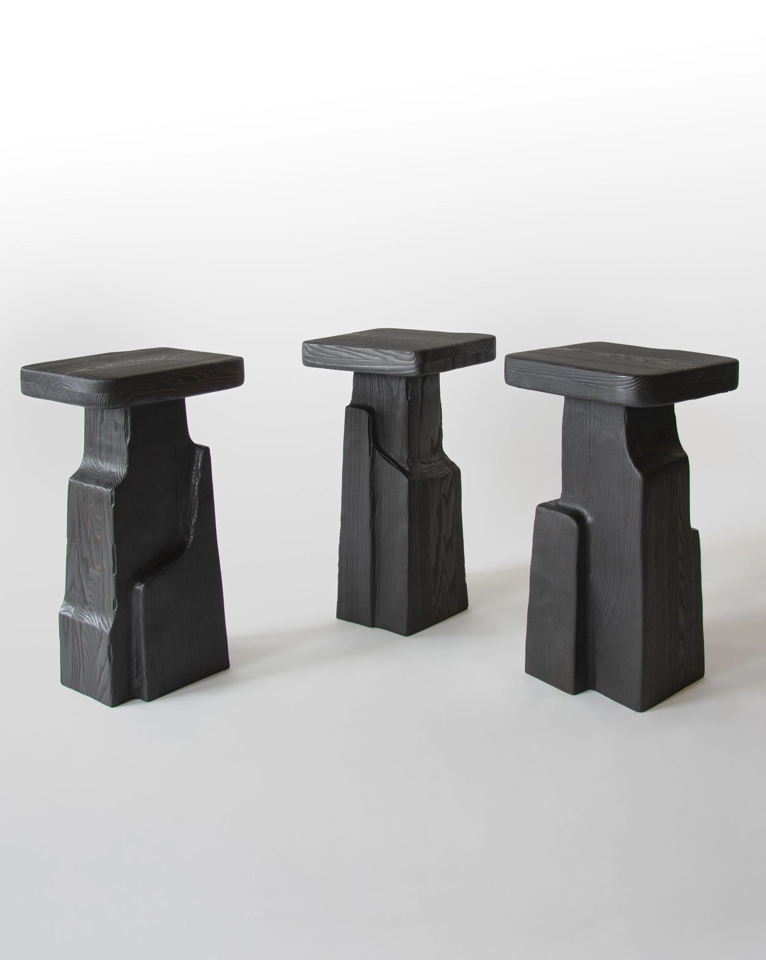 Three black wooden stools with rectangular seats and sculptural bases, arranged in a row on a white background.