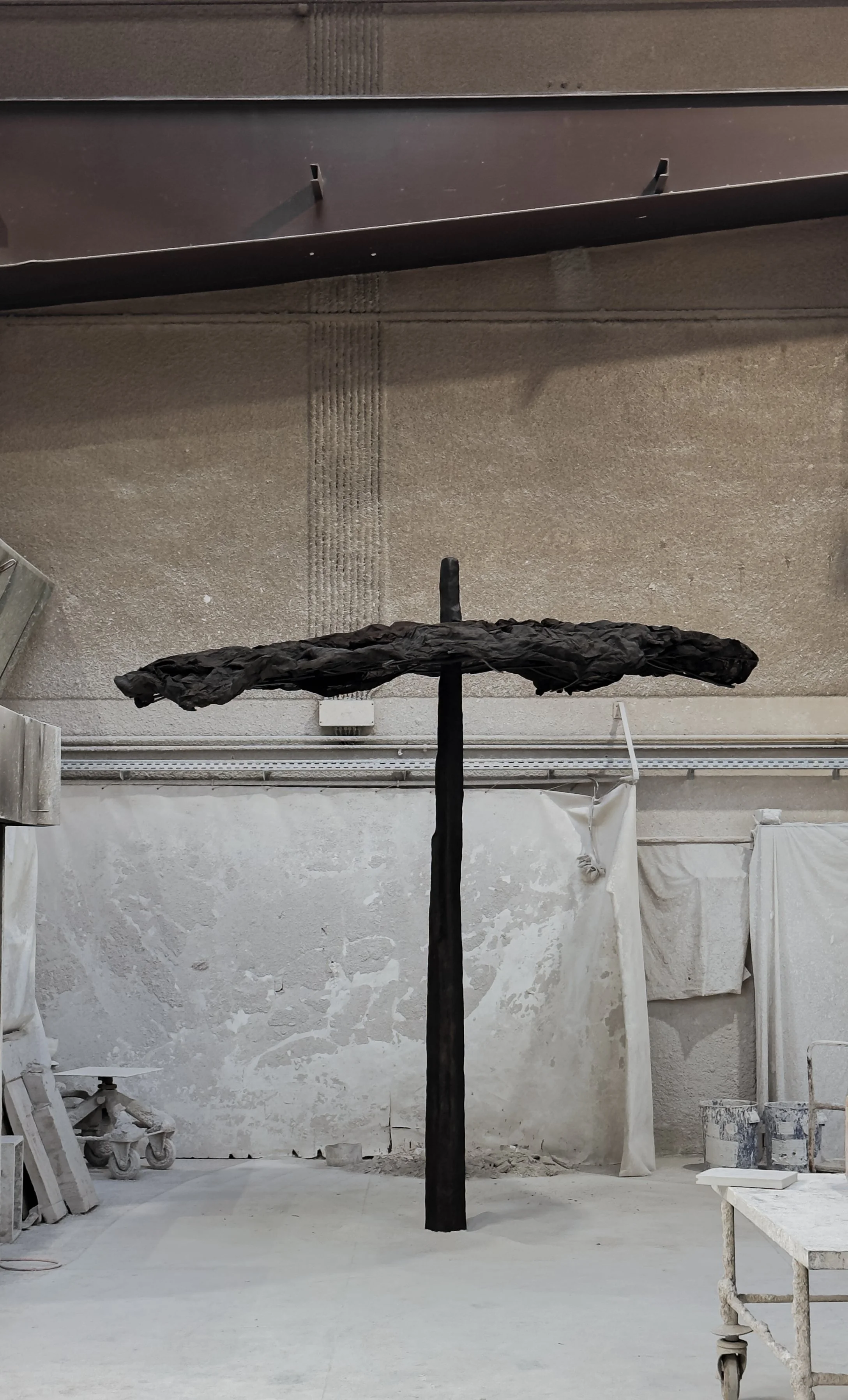 A large sculpture in an industrial studio, resembling a tall black pole with a wide, textured horizontal top, possibly made of paper or fabric, surrounded by dusty walls and workshop tools.