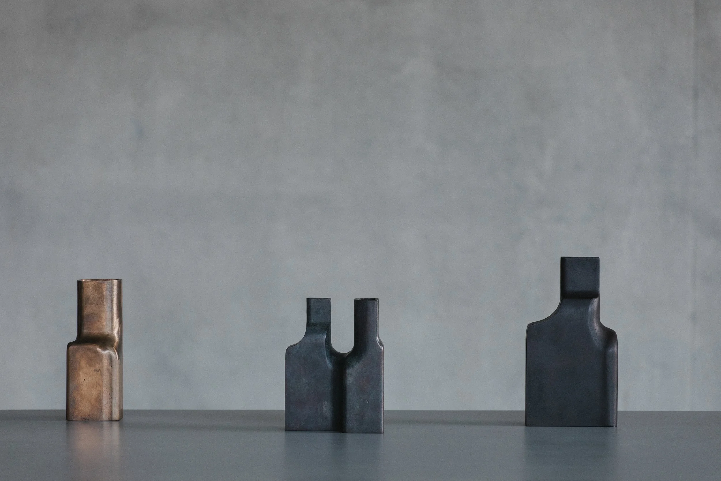 Three abstract stone sculptures of varying heights and colors on a gray surface with a plain gray wall background.