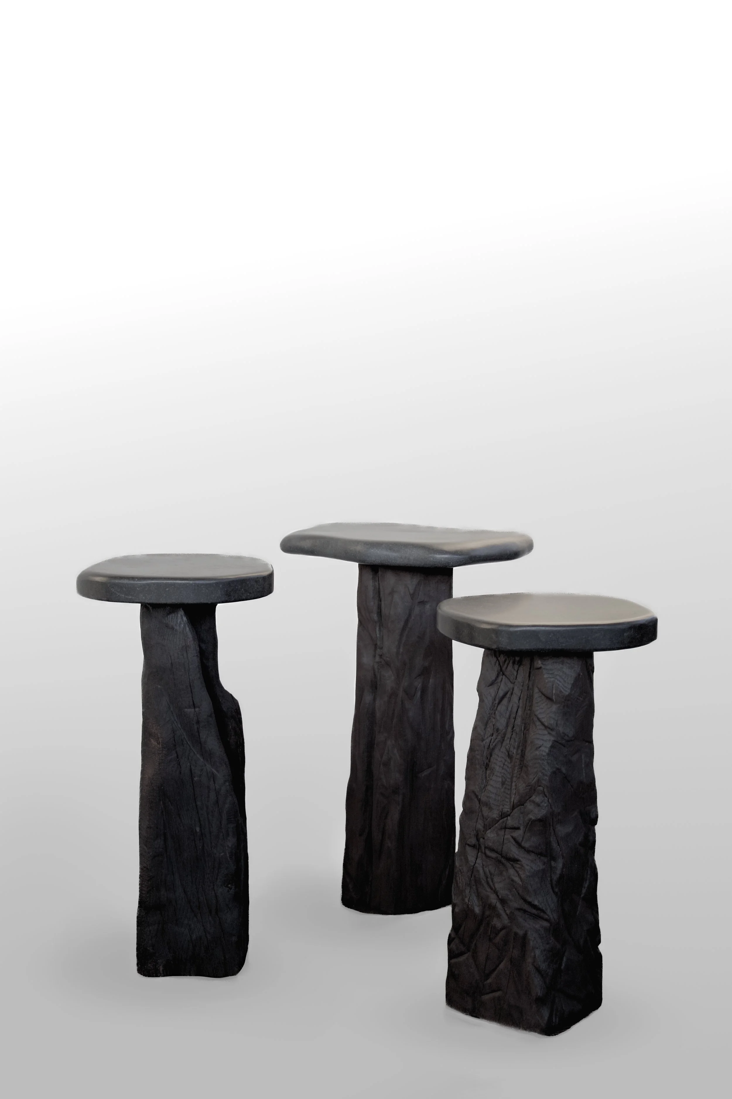 Three black wooden sidetables with square, flat, rounded tops, standing on rough, textured, irregularly shaped bases, displayed against a plain, light gray background.