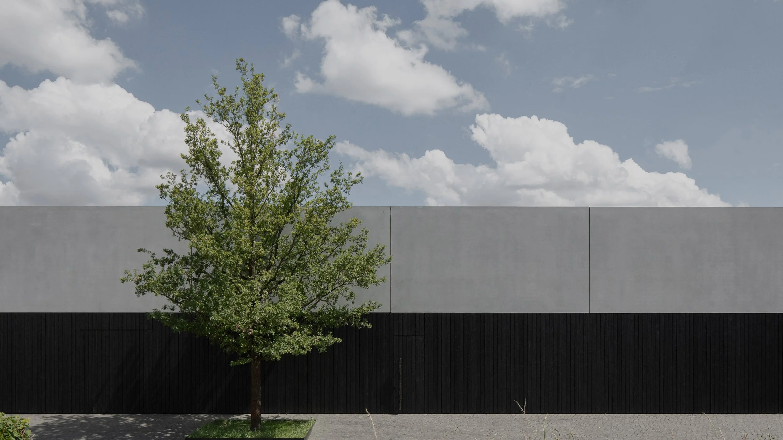 Minimalist building with a black lower section, a grey upper section, a tree to the left, and a partly cloudy sky.