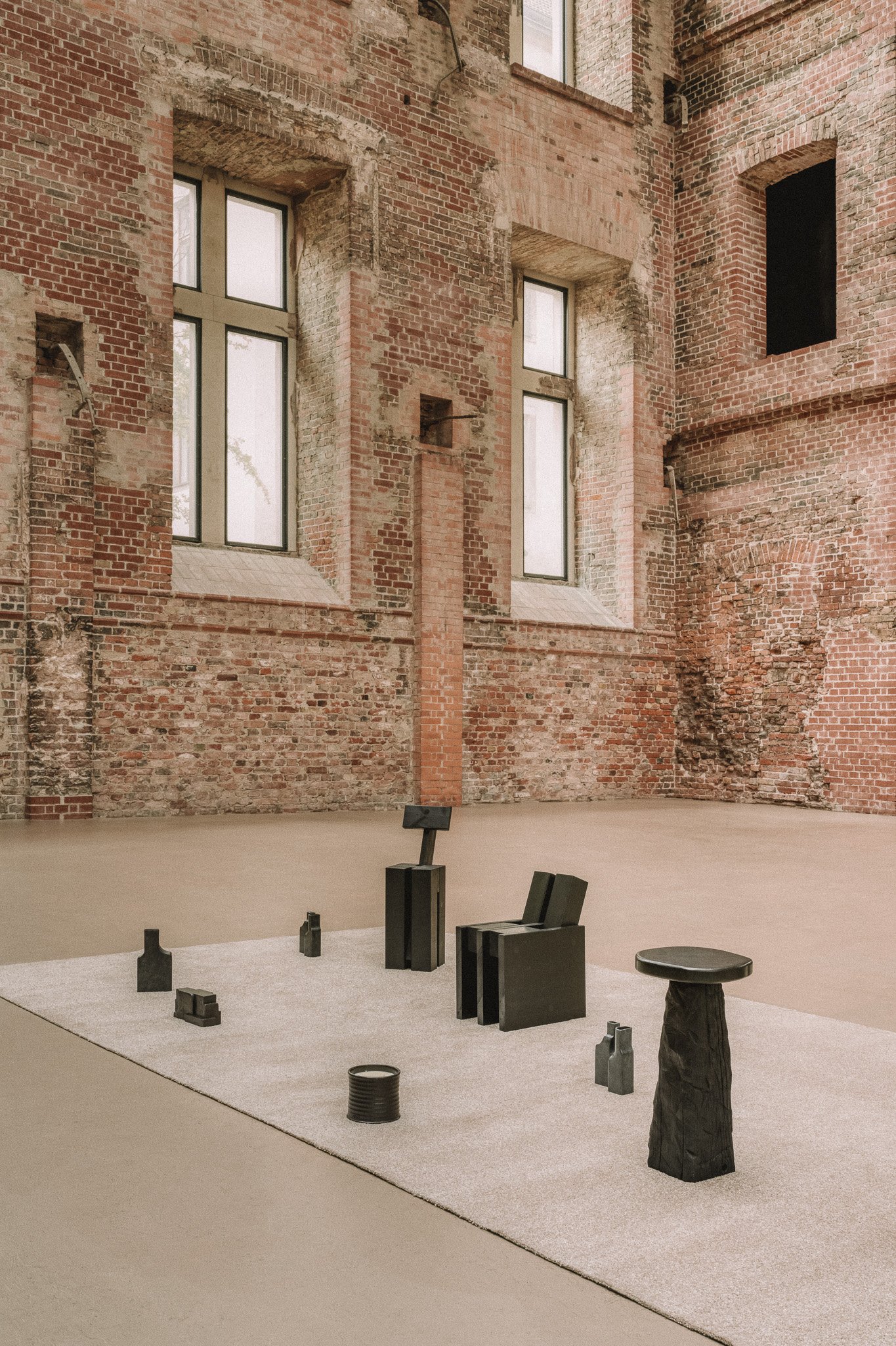 Art installation with black geometric sculptures on a light carpet in front of a exposed brick wall with large windows.