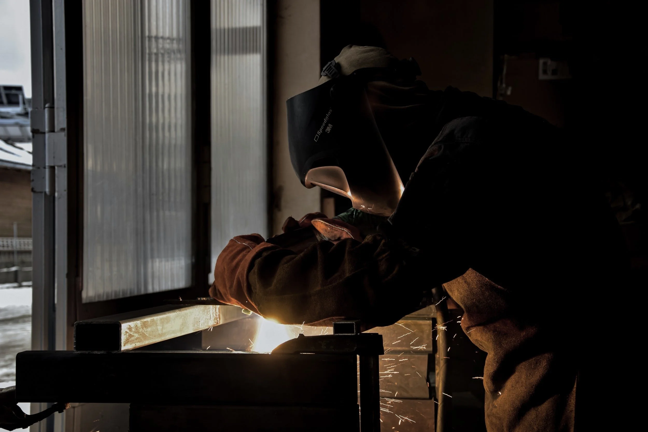 A worker welding metal in a workshop, wearing a protective welding helmet and gloves, with sparks flying from the welding process.