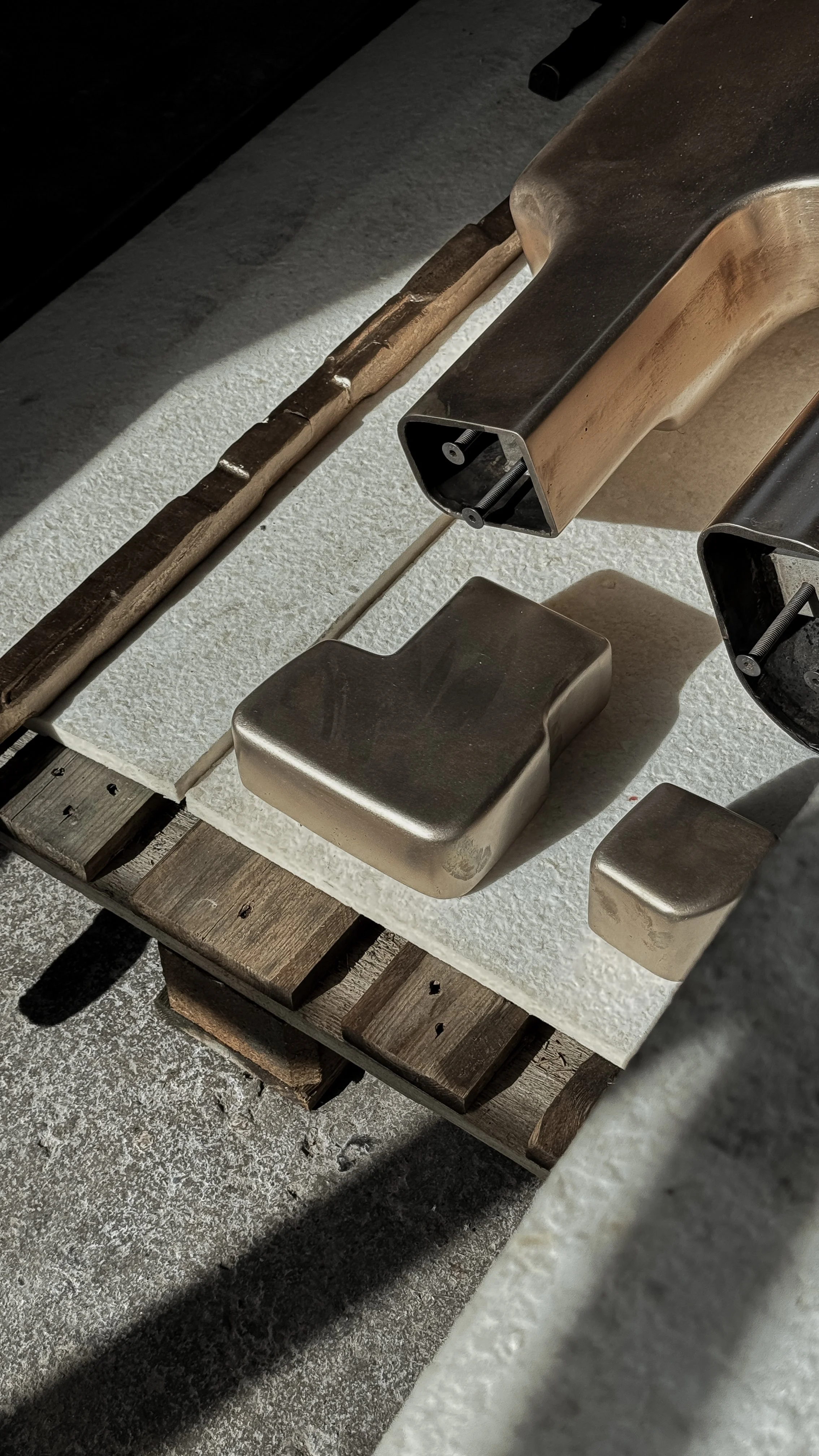 Close-up of a stainless steel countertop with two parts of a toaster, a white textured cutting board, and wood planks underneath, with sunlight casting shadows.