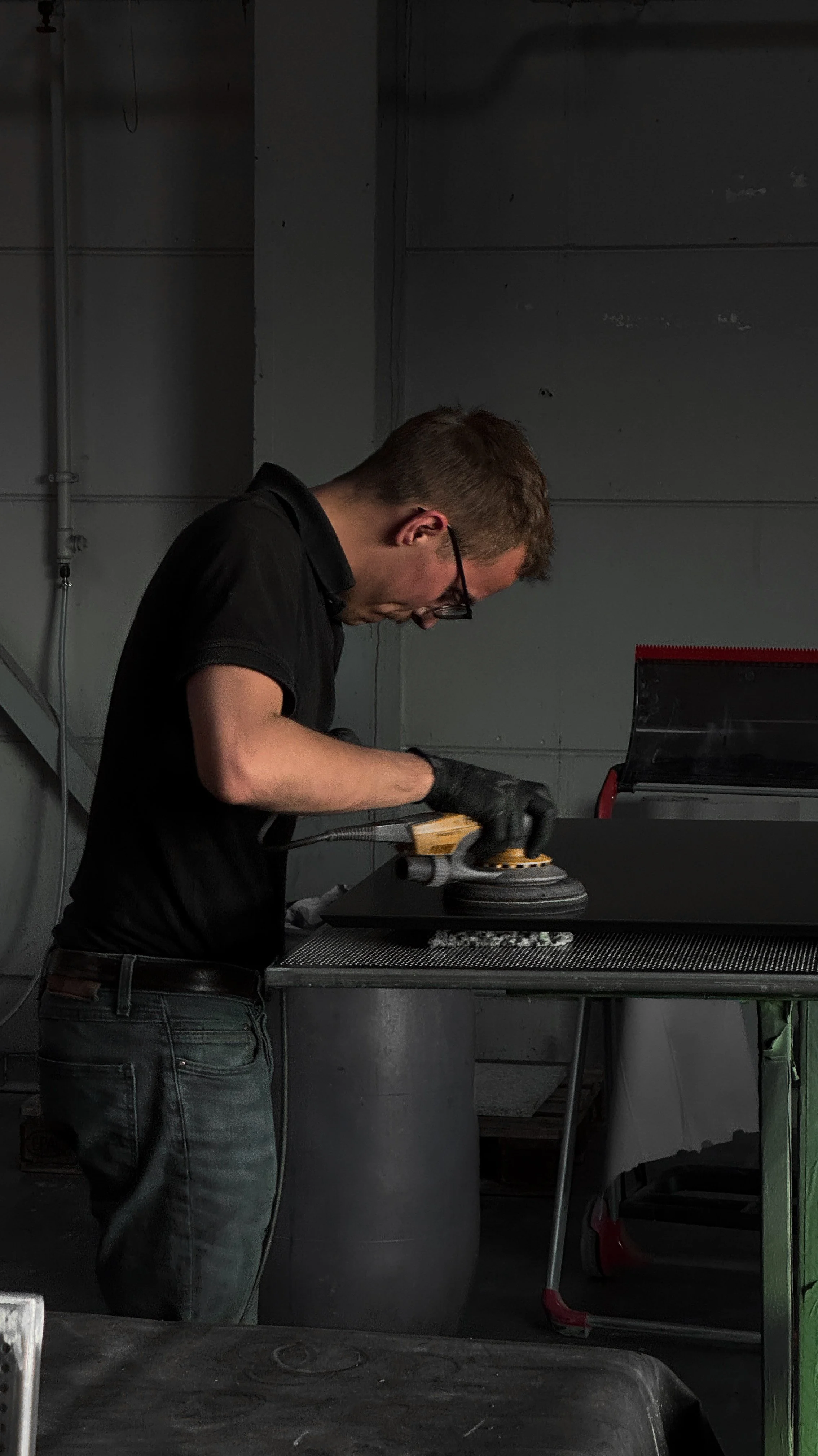 A man wearing glasses and a black shirt is polishing or sanding a black object on a workbench in a gray industrial or garage setting.